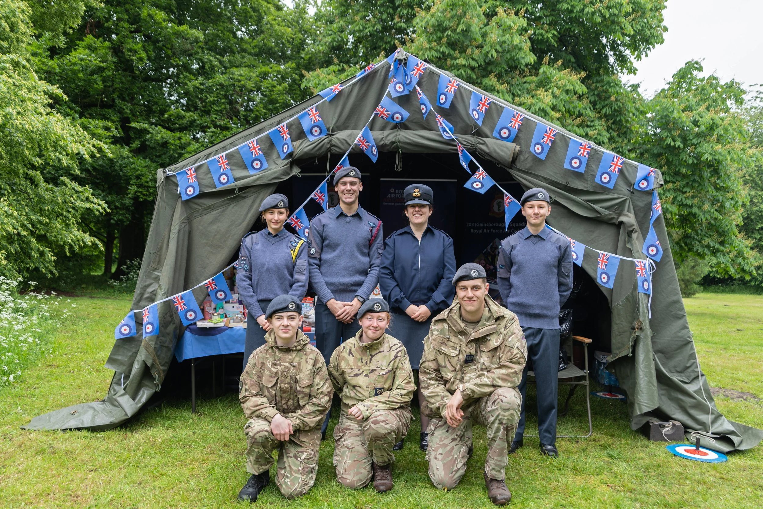 Group of young people in military and cadet uniforms standing and kneeling in front of a decorated green military tent outdoors, with British flags hanging on the tent, in a green park.