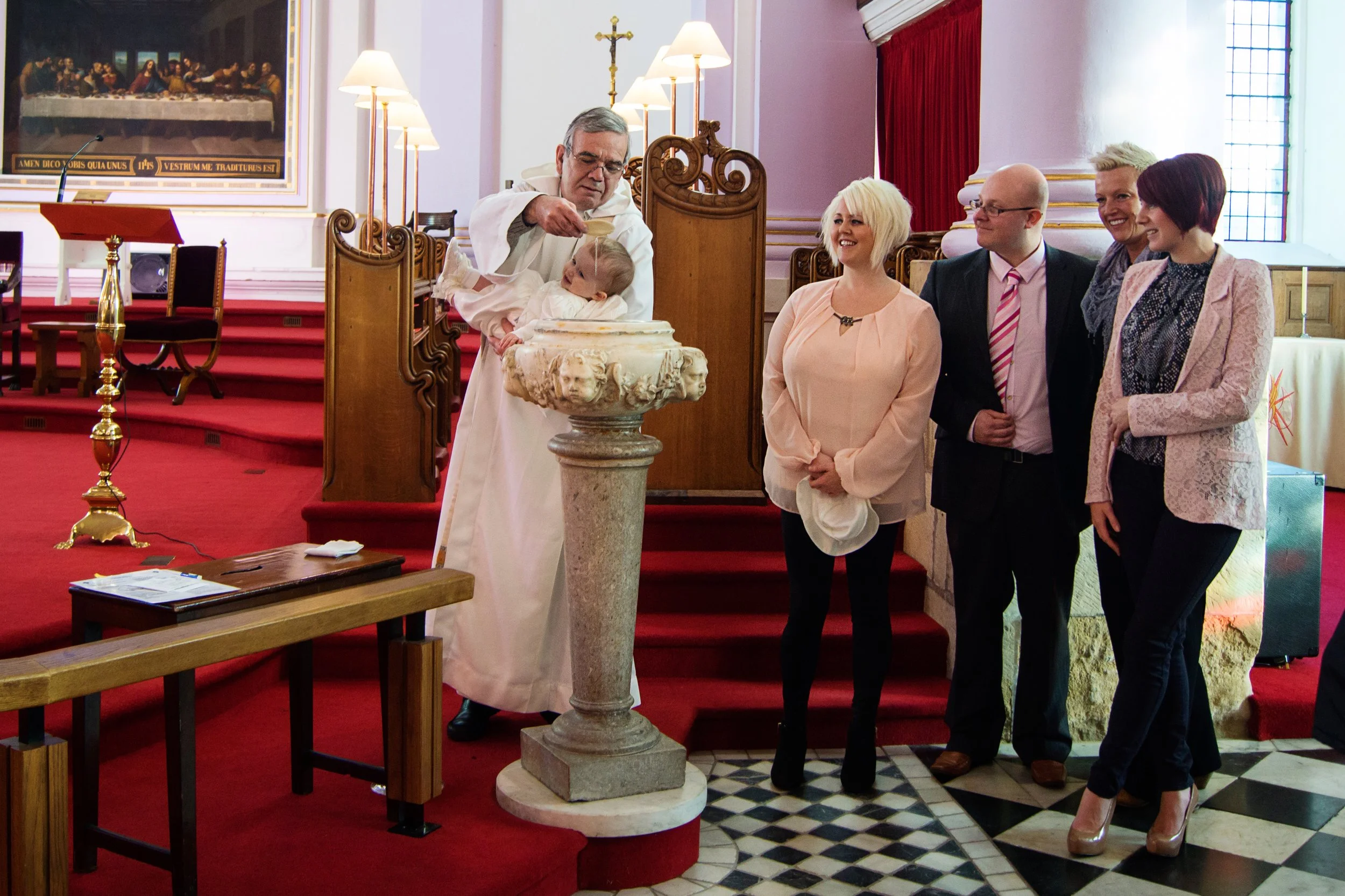 A priest baptizing a baby at a church, with three women and one man standing nearby, watching and smiling.