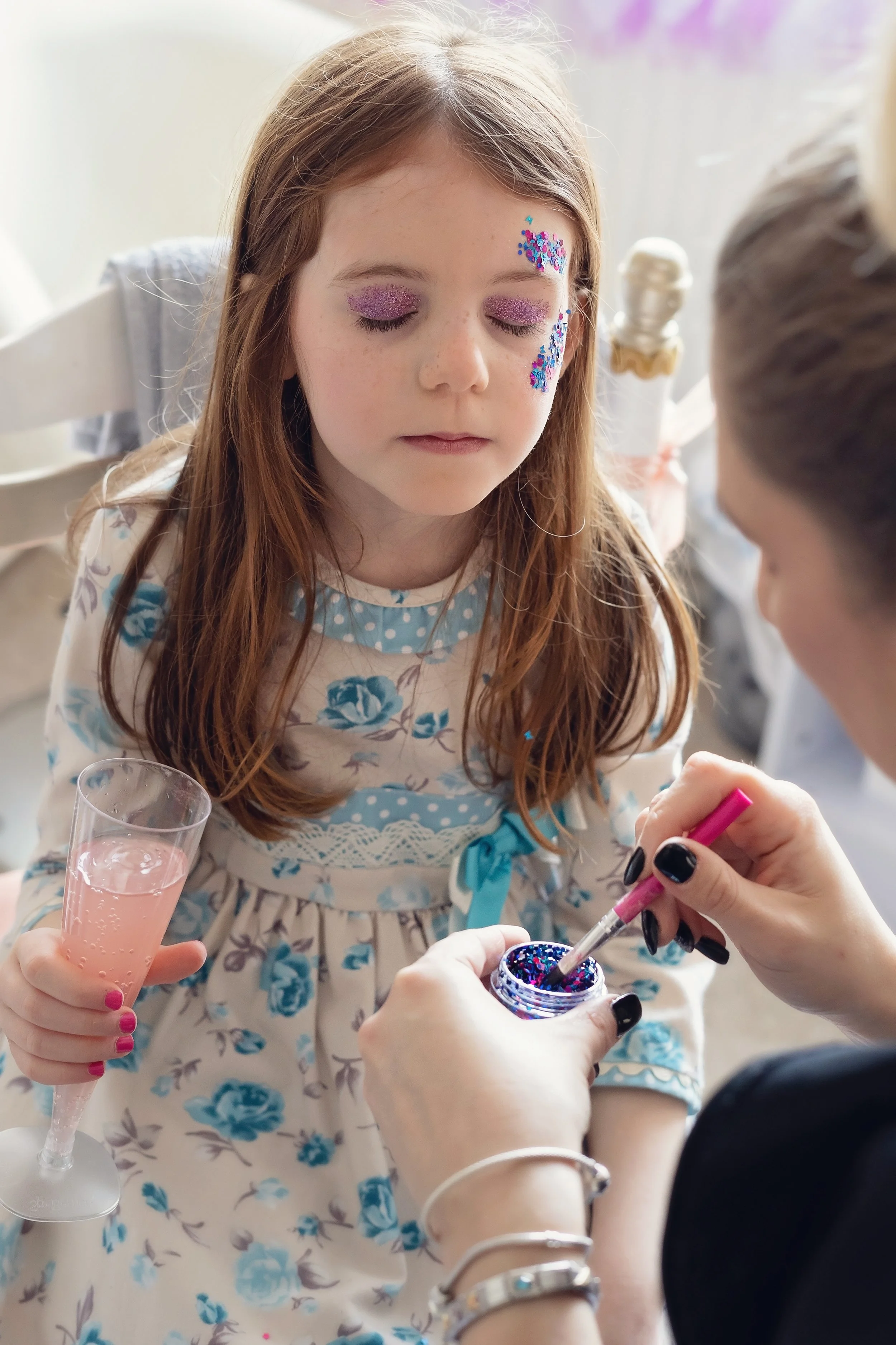 A young girl with long red hair and pink glittery makeup on her eyelids and face has her eyes closed as she gets glitter applied to her face. She holds a champagne flute filled with pink sparkling beverage, wearing a floral dress with blue accents. An adult applies glitter with a small brush.