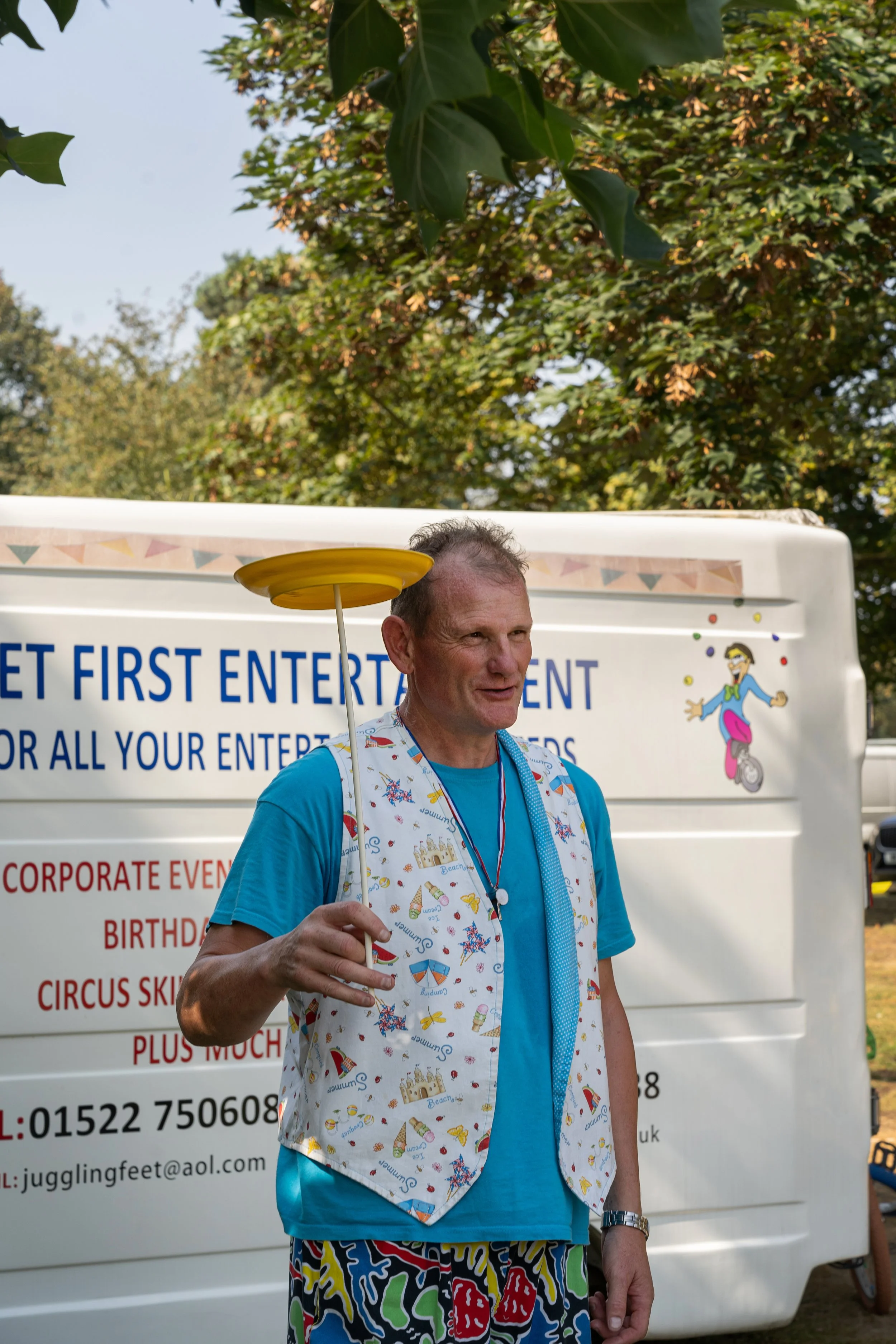 A man standing outdoors, balancing a yellow spinning plate on a stick in his hand, wearing a colorful vest and patterned shorts. There is a white vehicle with advertising behind him.