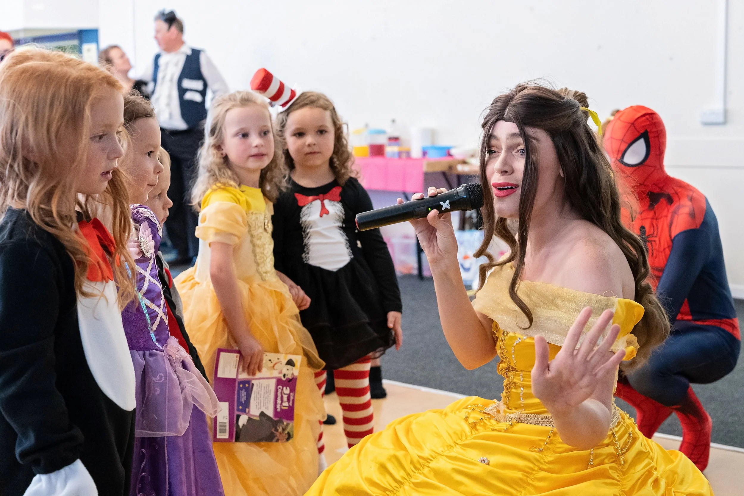 A woman dressed as Belle from 'Beauty and the Beast' singing into a microphone to a group of young girls dressed in princess costumes at a birthday party, with a person in a Spider-Man costume in the background.