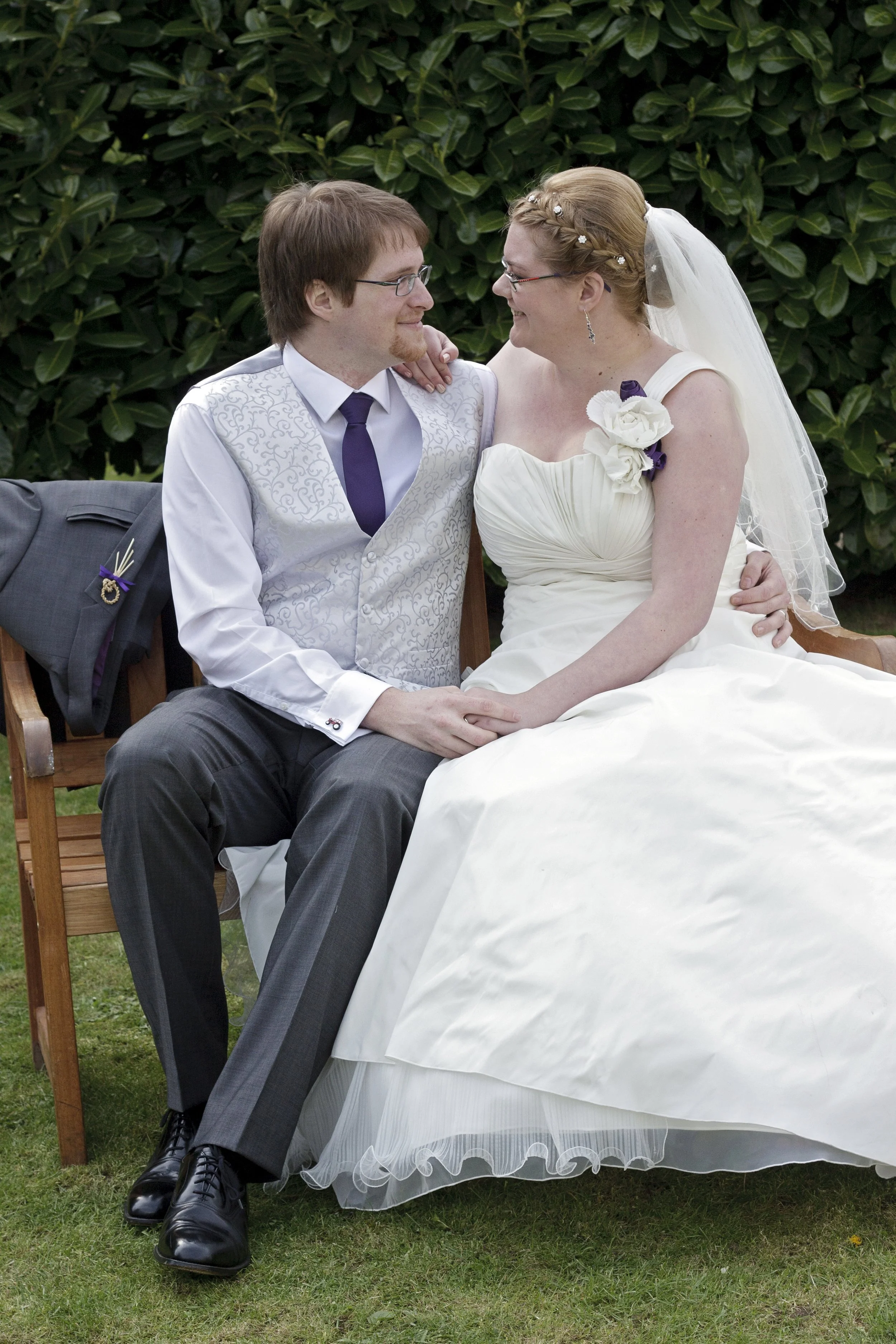 Bride and groom sat on bench, looking at each other and smiling.