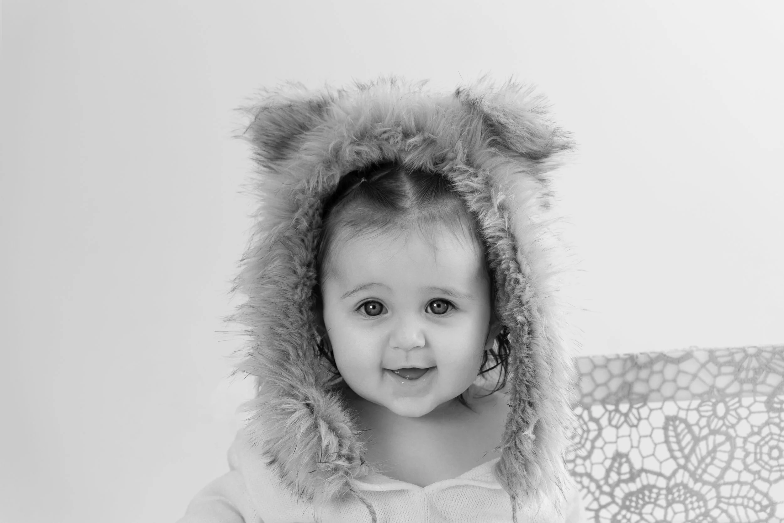 A smiling baby wearing a fluffy hat resembling a lion's mane, sitting in front of a plain background with a woven patterned object in the background.