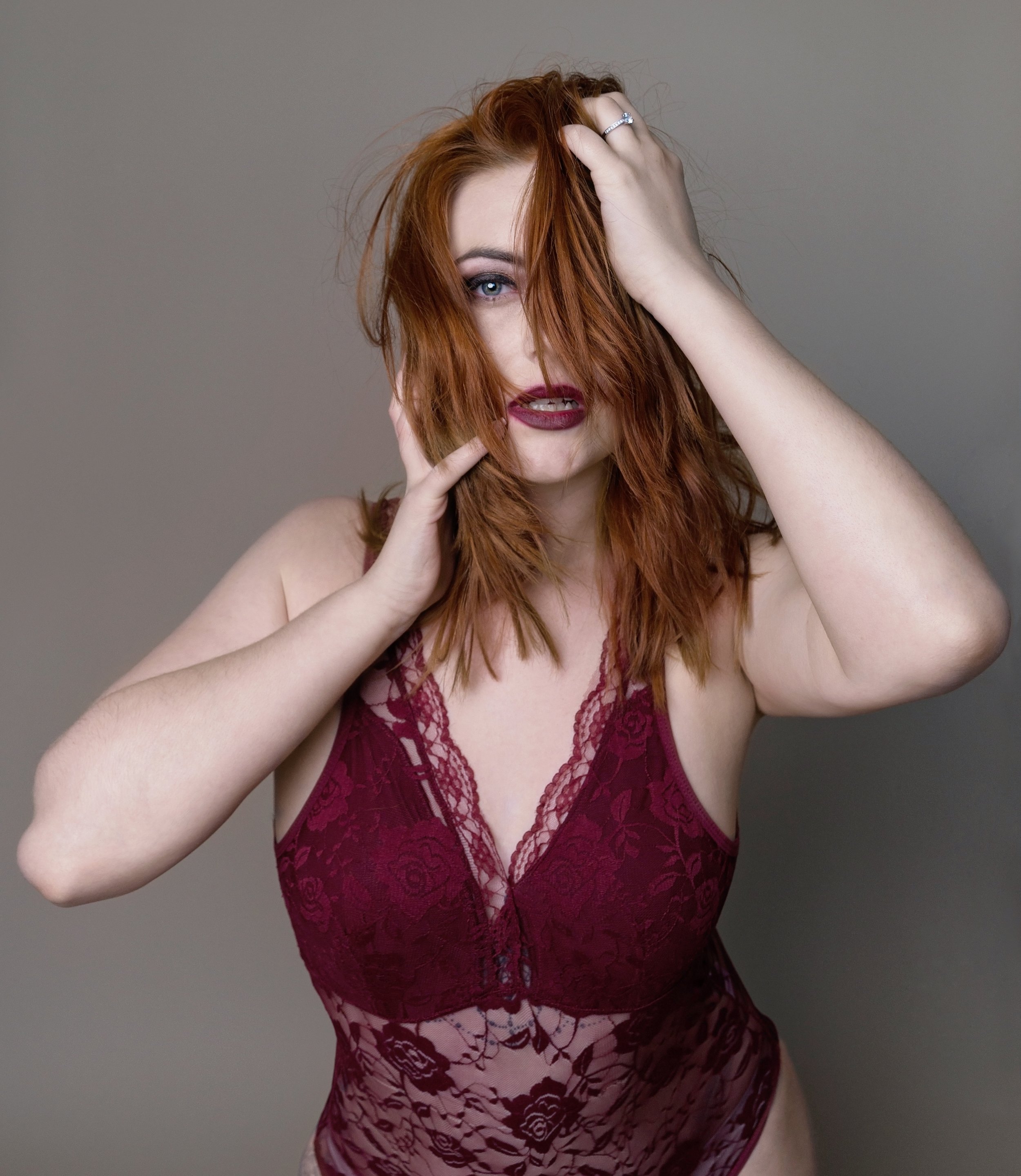 A woman with reddish hair and blue eyes wearing a burgundy lace bodysuit, touching her face and hair, with a neutral gray background.