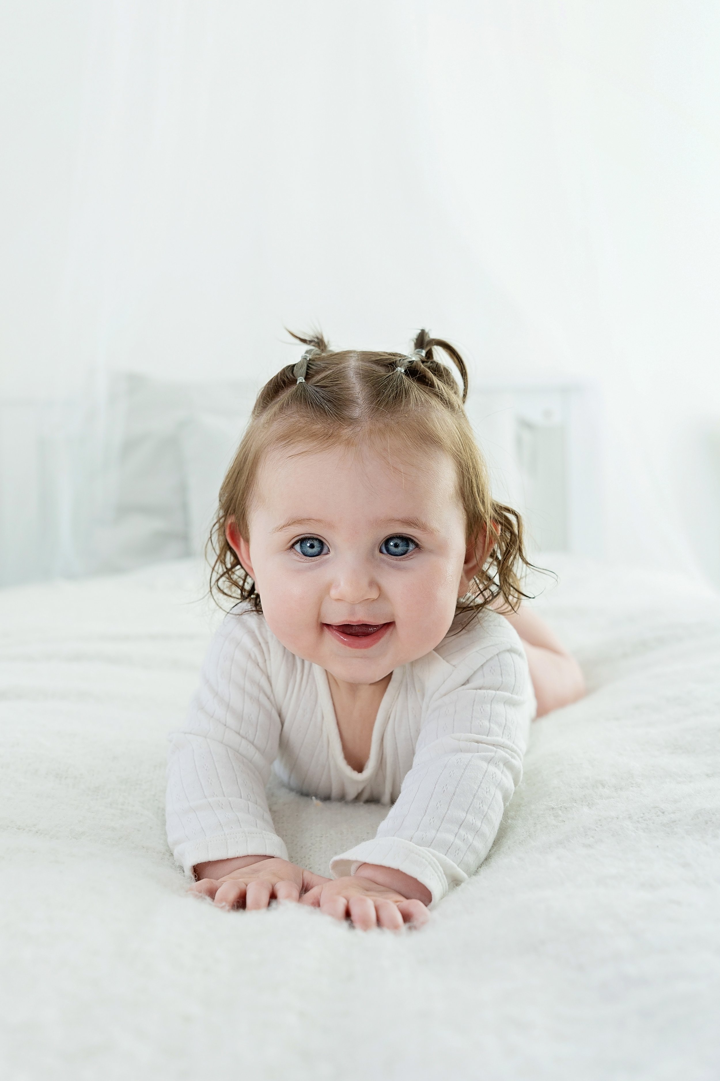 A smiling toddler with blue eyes and light brown hair, styled with small pigtails, crawling on a white bed in a bright room.