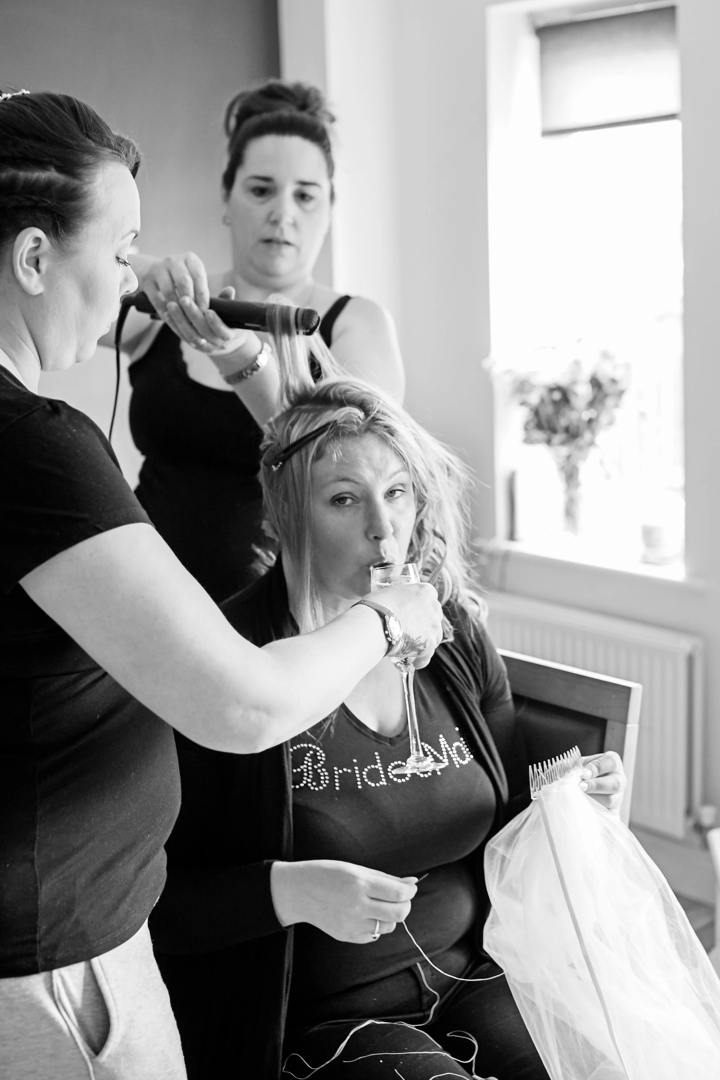 Bridesmaid multitasking, she is sewing a vail, getting her hair done and another bridesmaid is holding champagne for her to drink - Black and white photo.