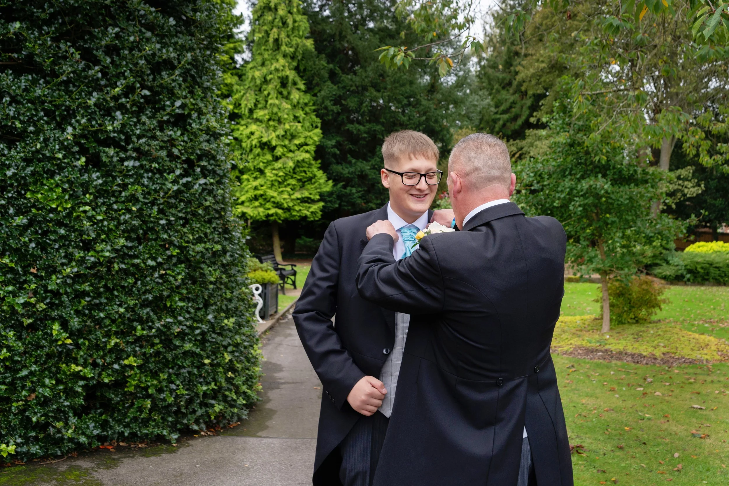 Color photo outdoors of father doing up late teenage sons colar, with trees in background