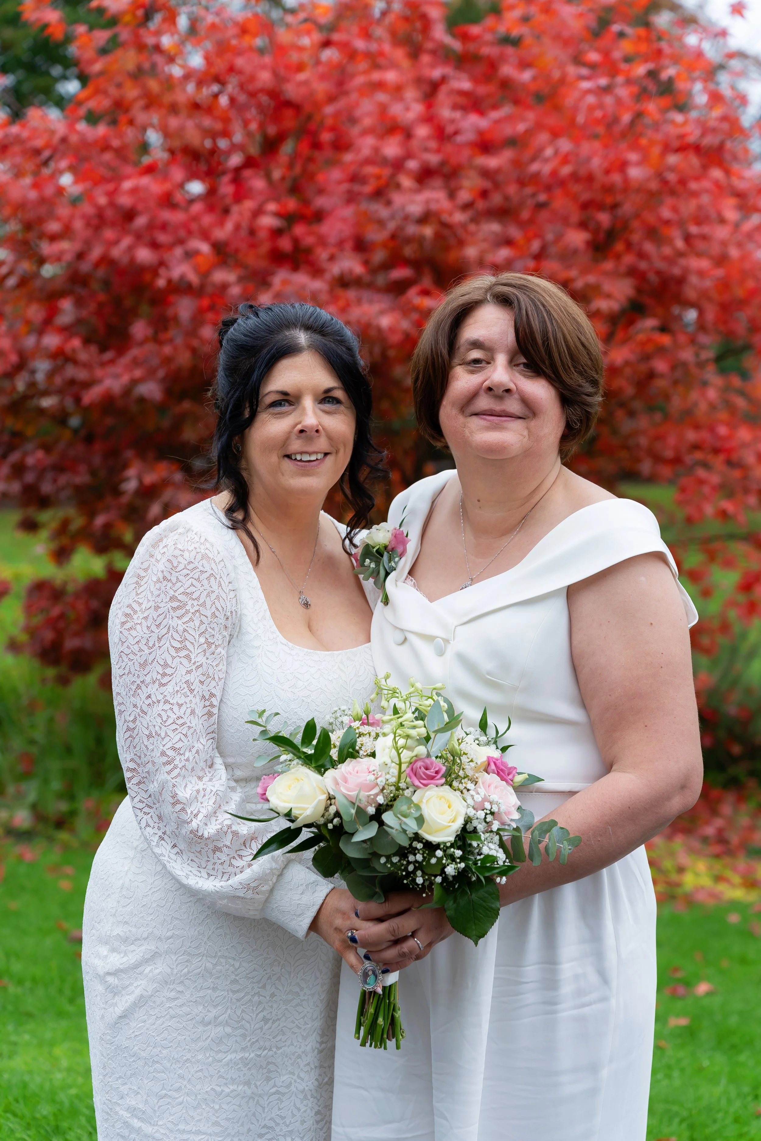 Lesbian couple with Red tree behind them, both looking at camera smiling, holding bouquet of flowers at Gainsborough Registry office.