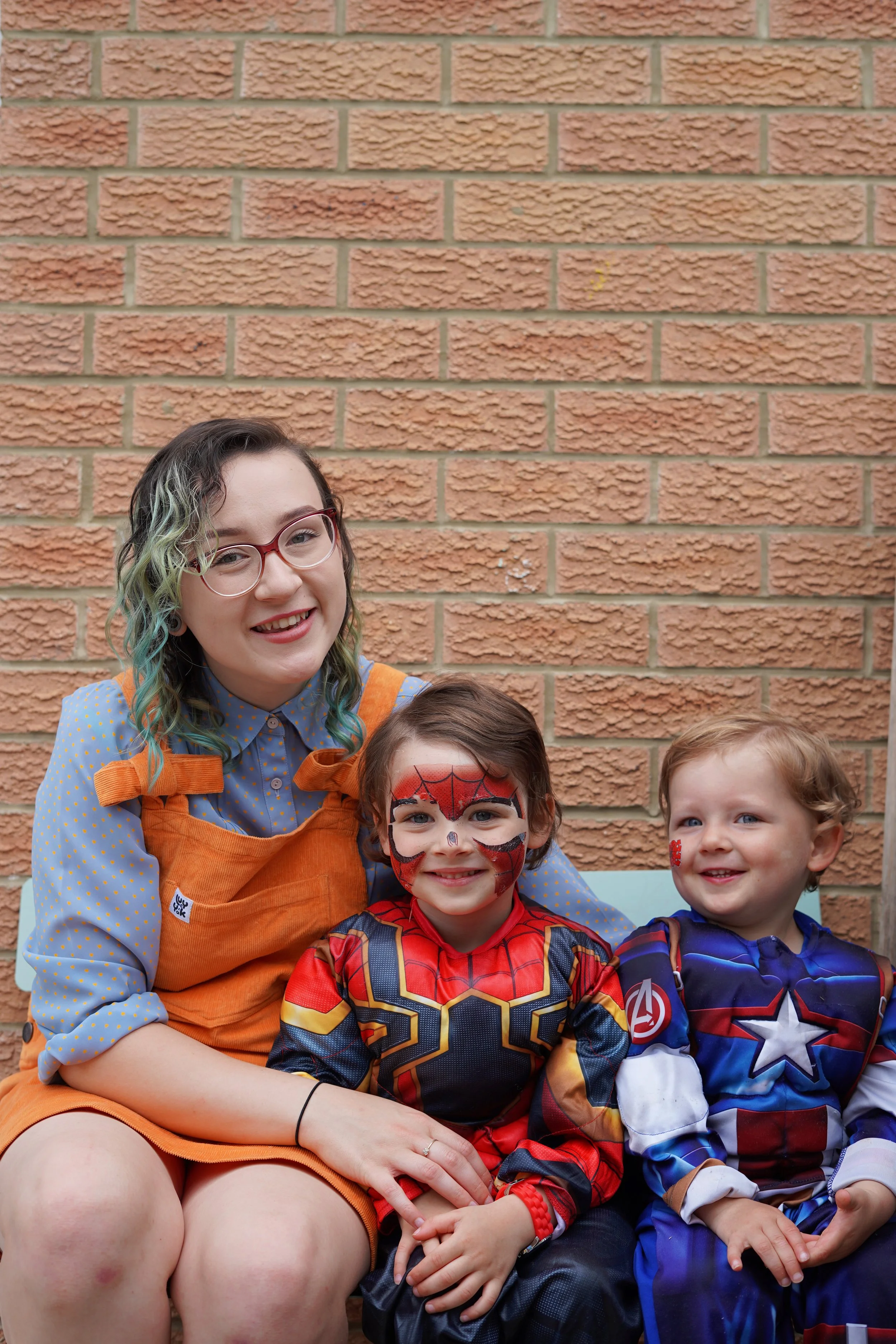A young woman and two children dressed in superhero costumes sitting against a brick wall.
