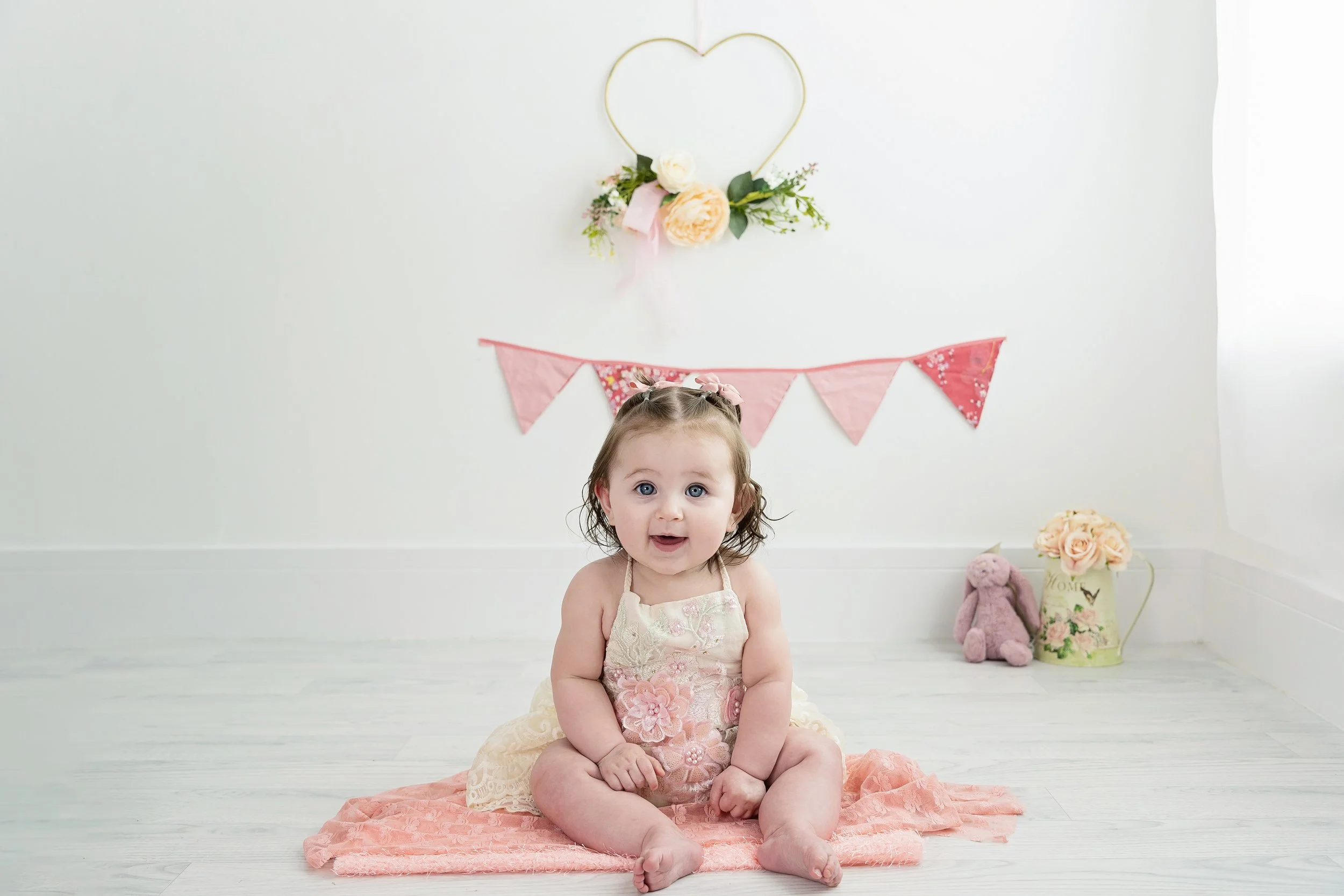 A young girl with curly brown hair, blue eyes, and a pink bow, sitting on a pink blanket on a white wooden floor in a decorated room with pink bunting, a heart-shaped wall decoration, and a vase of flowers in the background.