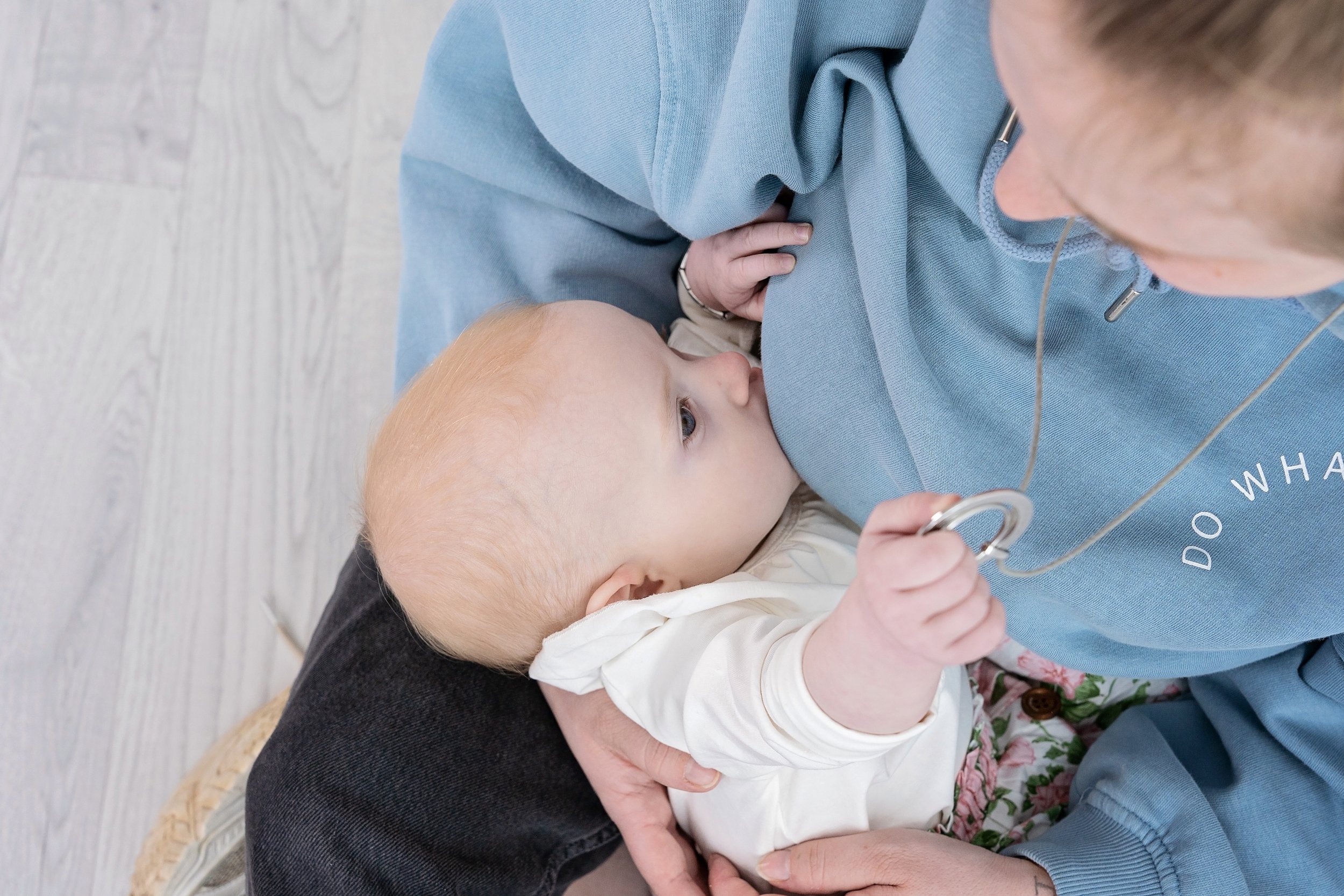 A young child with blonde hair nursing on a woman wearing a blue hoodie, with the woman holding a stethoscope.