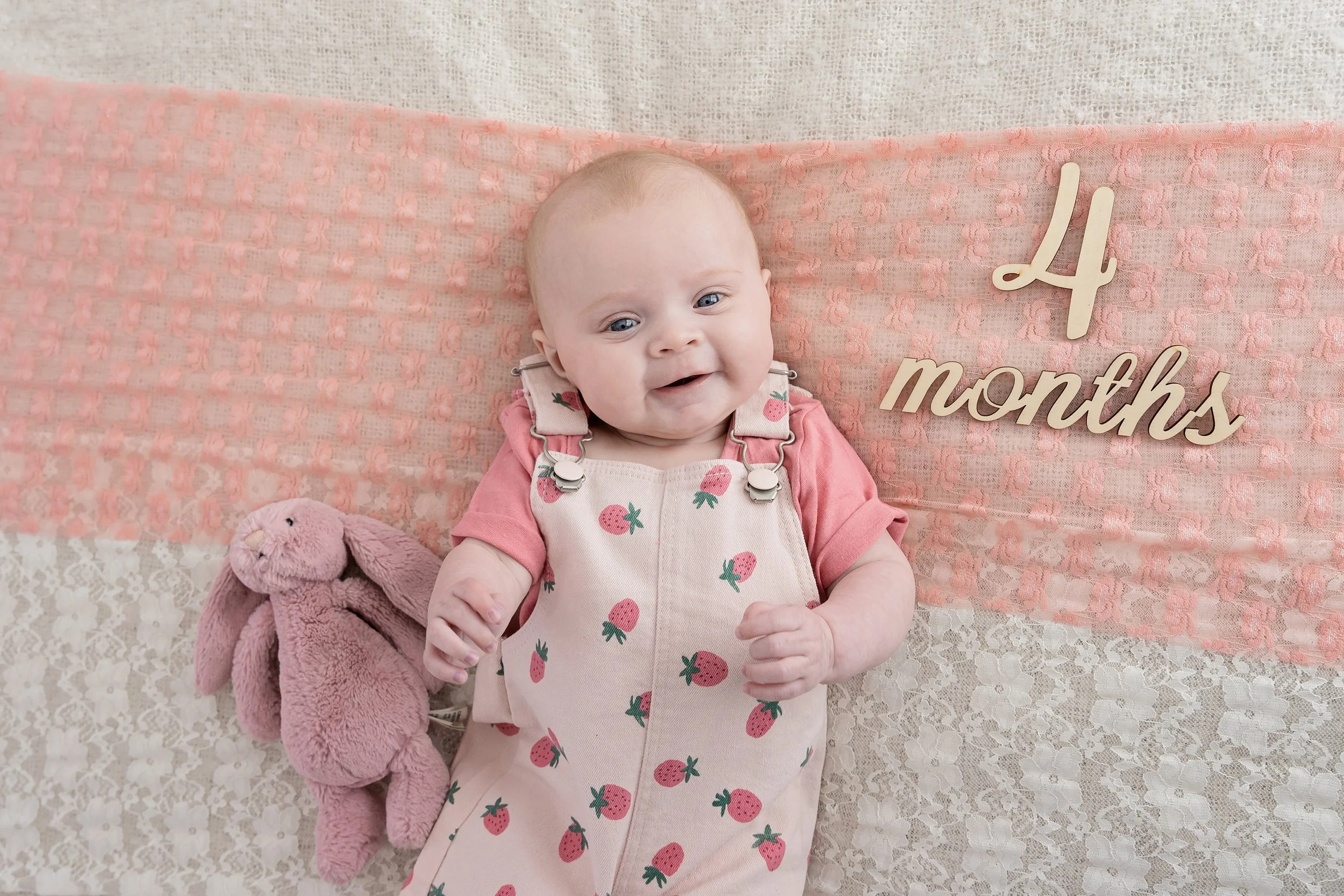 A four-month-old baby girl with light hair and blue eyes, lying on a lace blanket with pink lace and a wooden sign that reads '4 months'. The baby is smiling and wearing a pink shirt with pink strawberry print over pink overalls. A pink stuffed bunny is next to her.