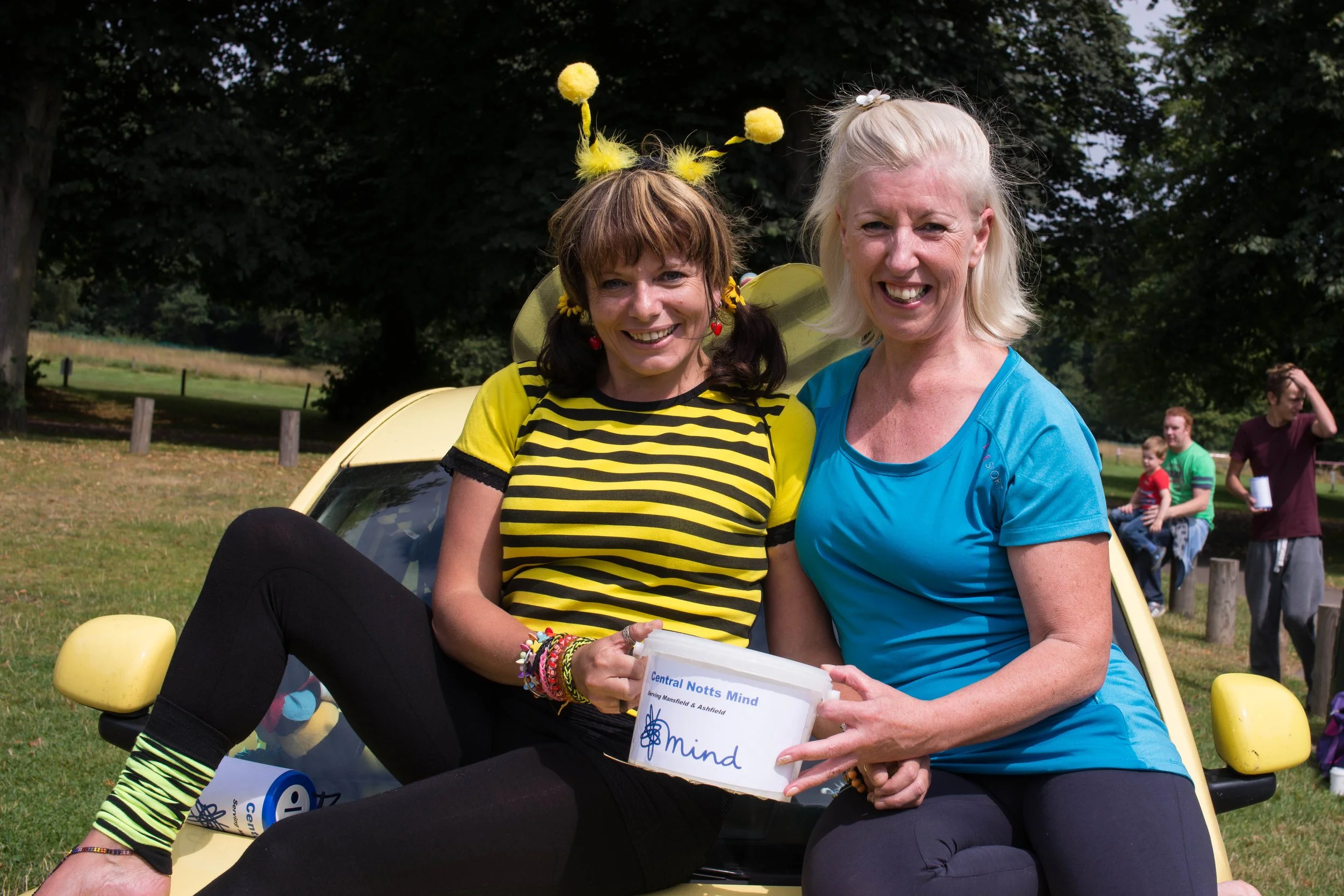 Two women sitting on a yellow car outdoors, smiling and holding a donation box for a charity called 'Central Notts Mind,' with people in the background enjoying a sunny day in a park.