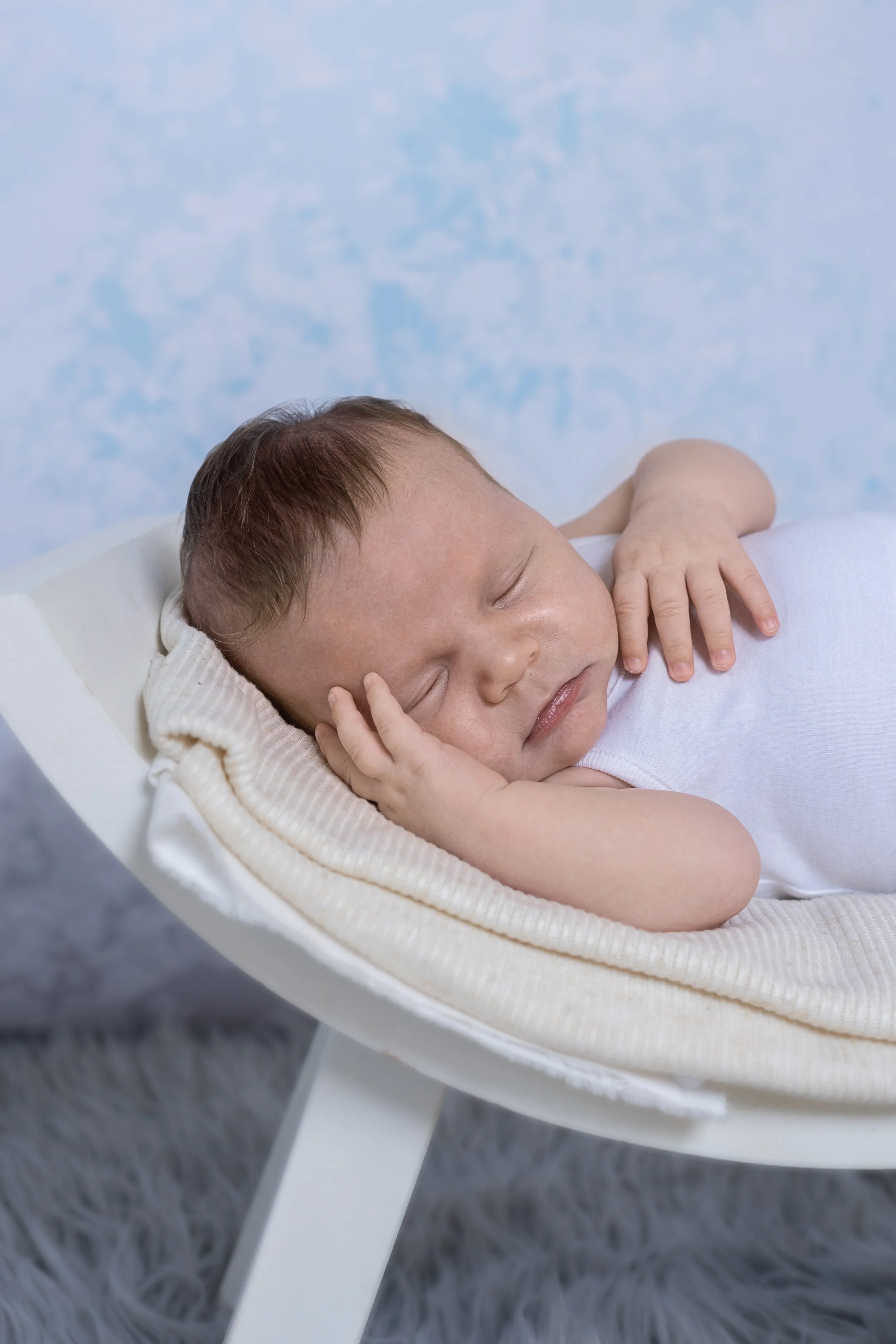 A sleeping baby with brown hair lying on a white cushioned cradle with a beige border, wearing a white sleeveless shirt, resting peacefully with one hand on his face and the other on his chest.