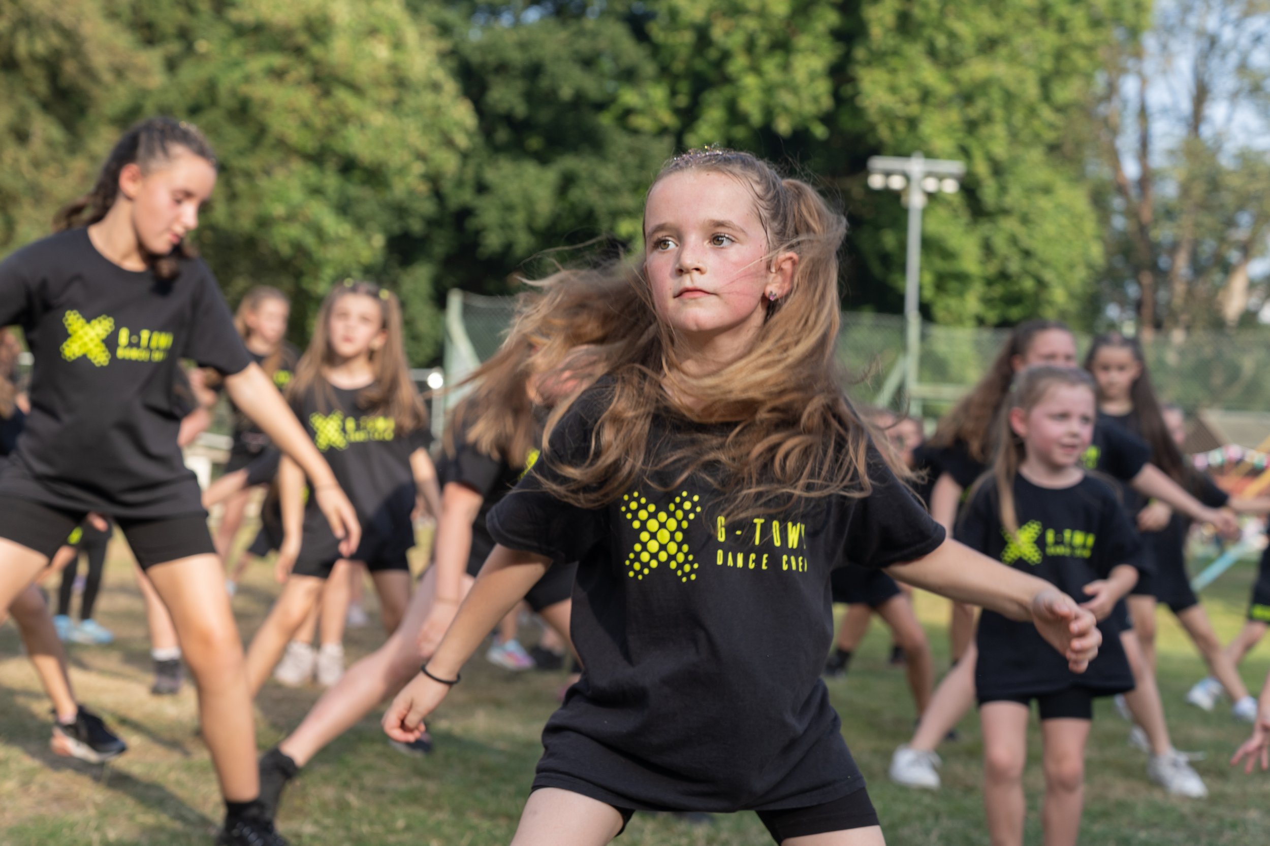 A group of young girls dancing outdoors on a grassy field, wearing black T-shirts with neon yellow graphics and text, with trees and a fence in the background.