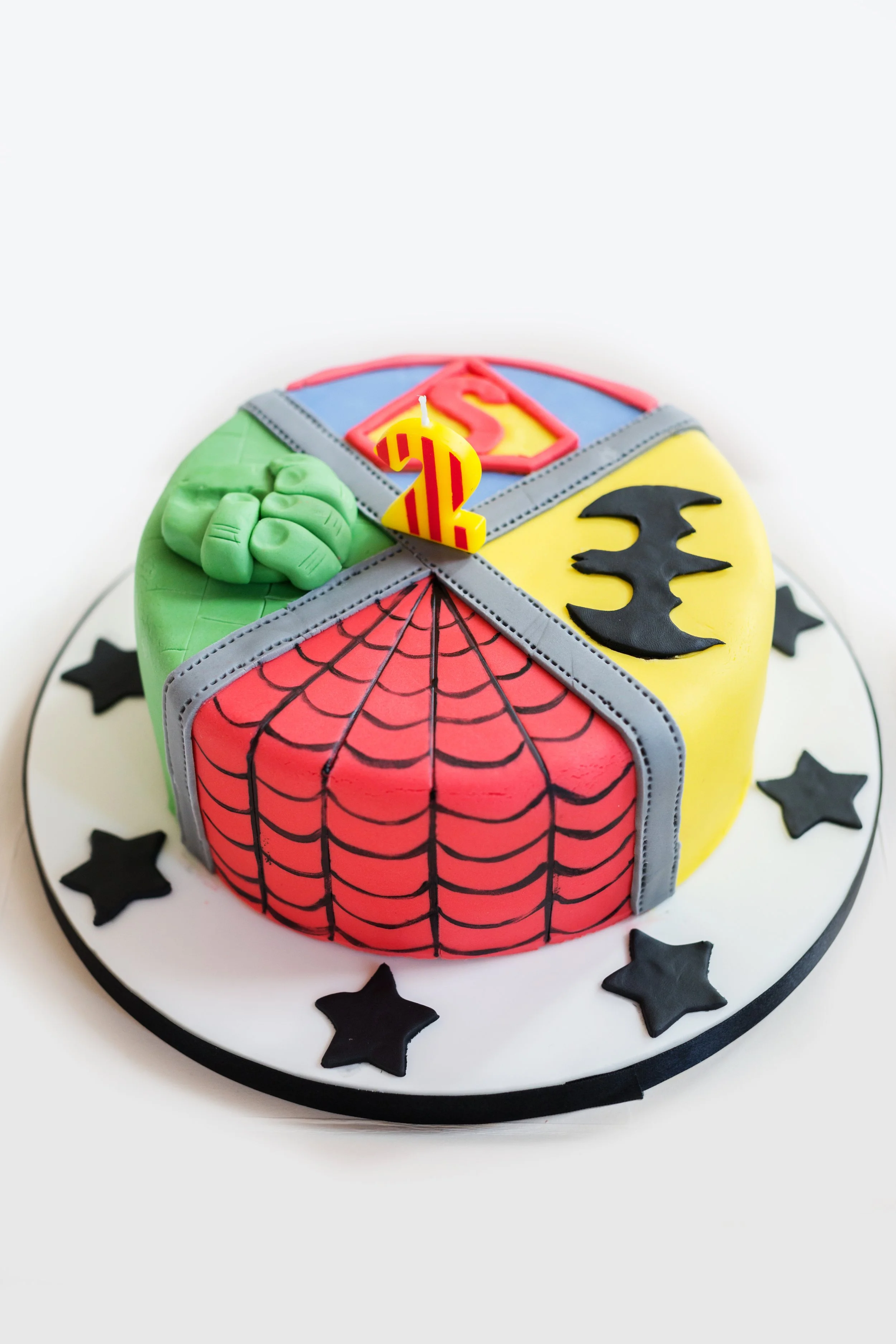 A round Superman-themed birthday cake decorated with superhero symbols and a Superman logo candle.