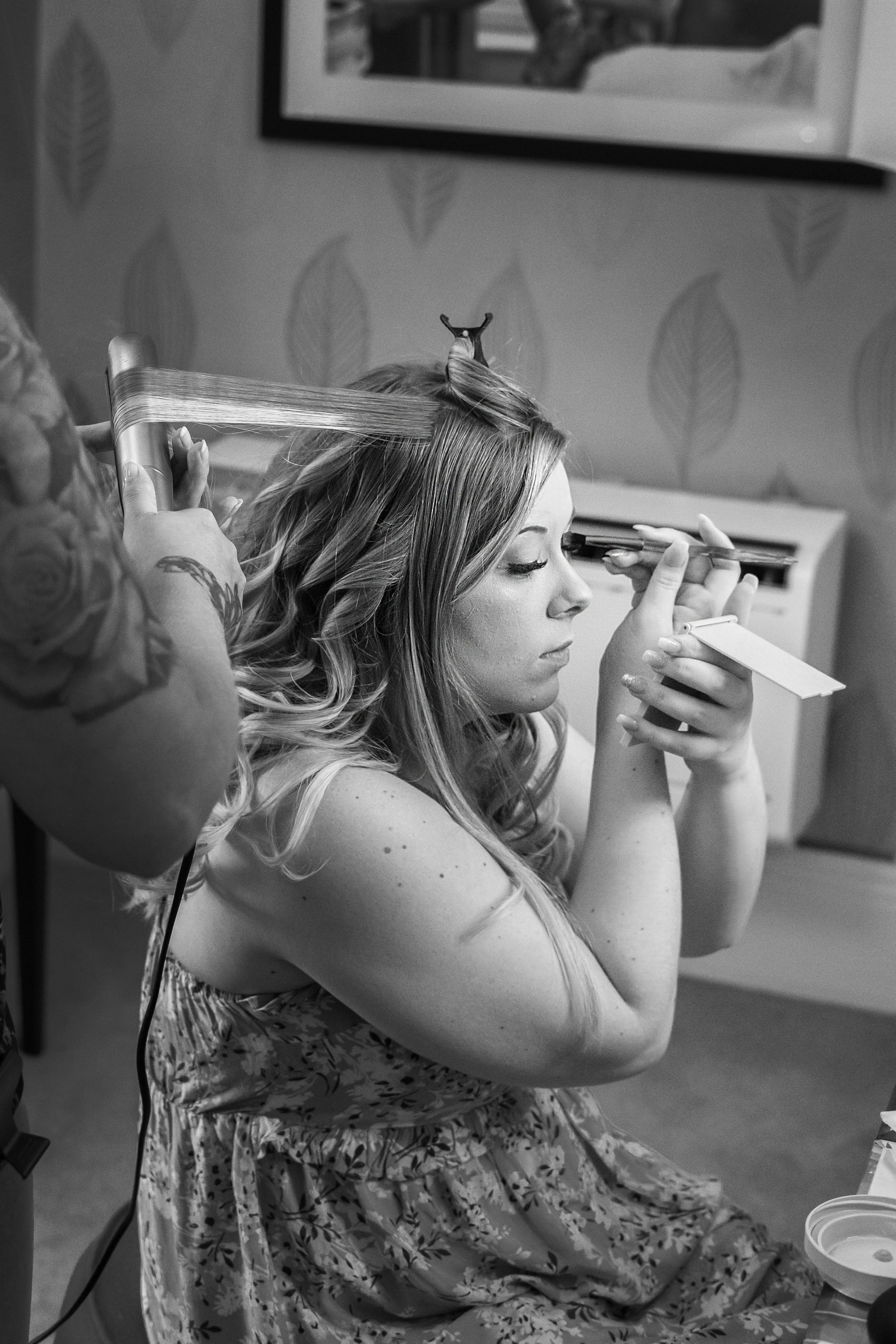 A woman with long, wavy hair is having her makeup done while a stylist is working on her hair, using a curling iron. She is sitting and looking into a mirror, with a makeup palette in her hand.