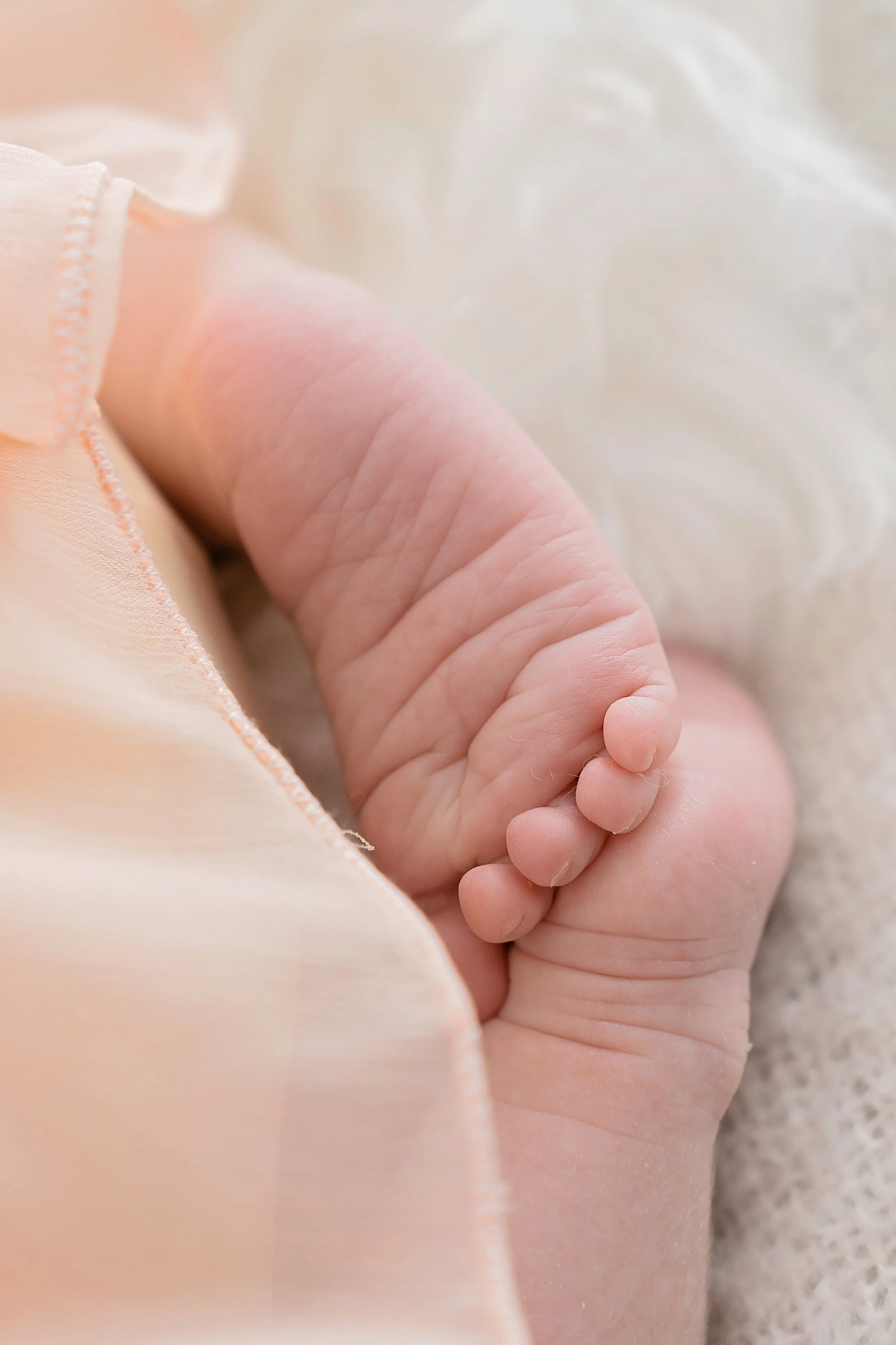 Close-up of a baby's tiny hand gripping an adult's finger, with soft, pastel-colored clothing and blanket in the background.