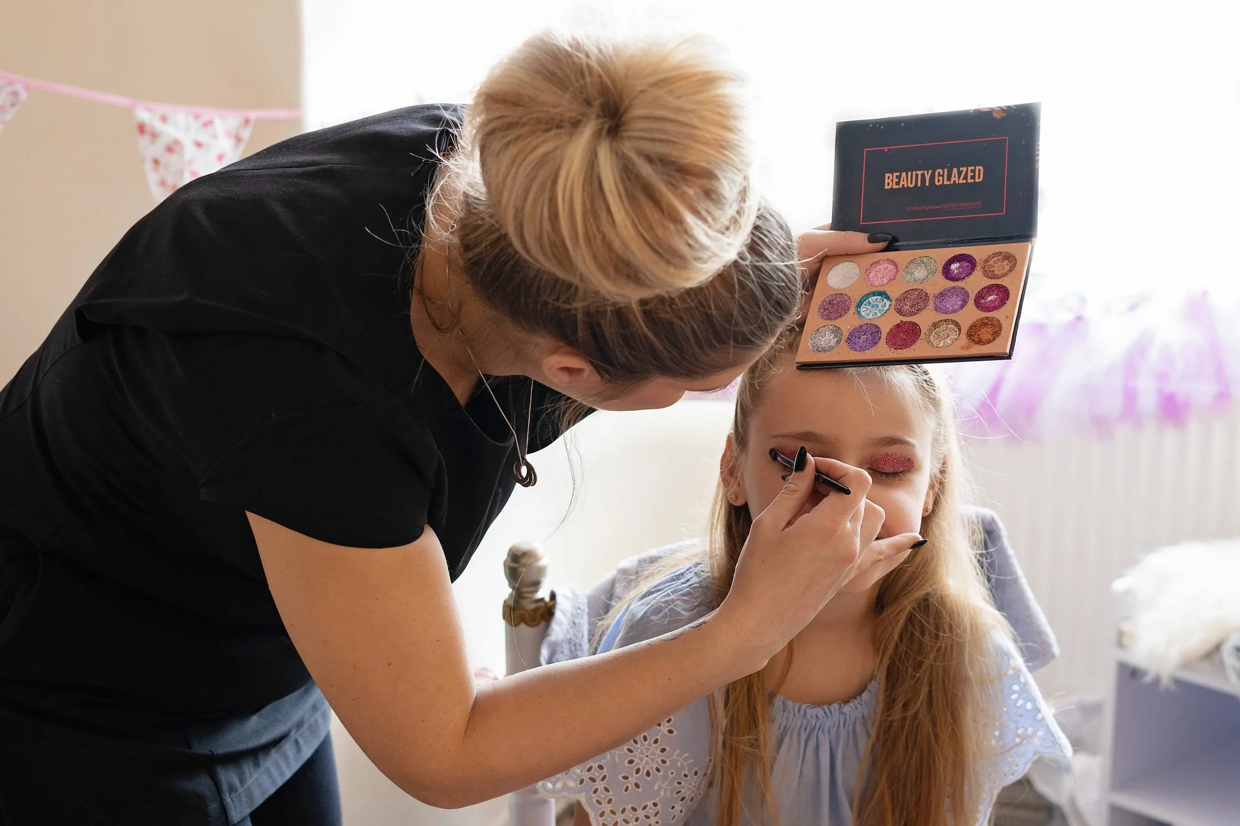 Adult woman applying eyeshadow to a young girl using a makeup palette labeled 'Beauty Glazed' in a bright room.