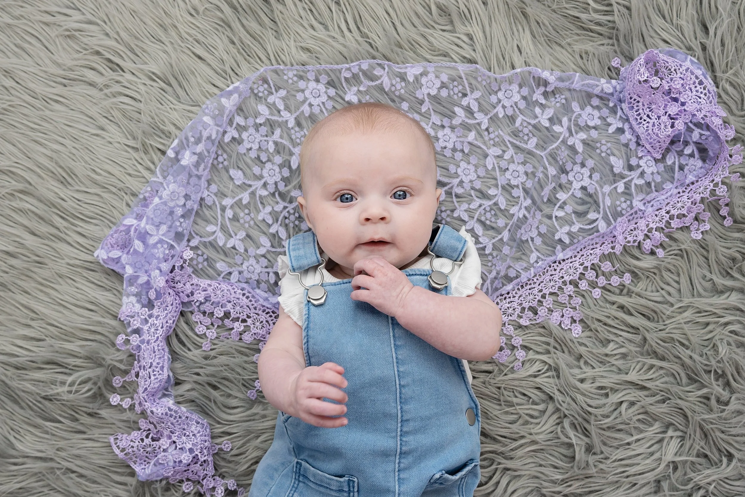 A baby with blue eyes lying on a gray, fluffy surface, surrounded by a purple lace cloth, wearing a white shirt and denim overalls.