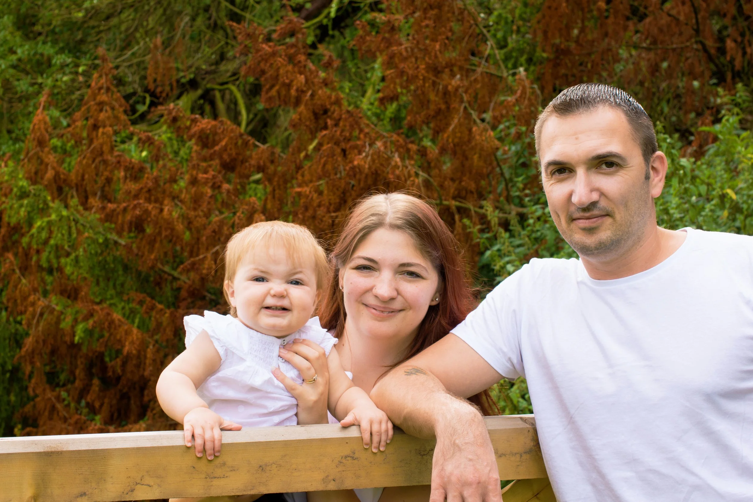 Family of three, a man, woman, and young girl, outdoors in front of trees with brown and green foliage, leaning on a wooden railing and smiling.