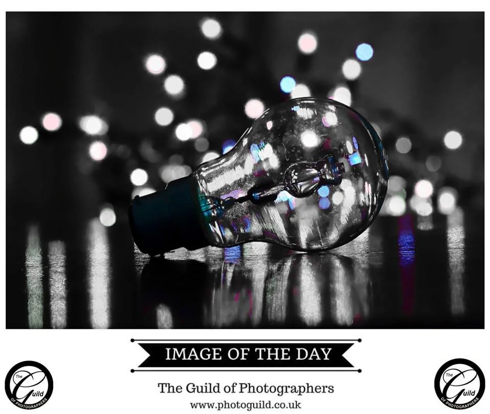 A clear glass light bulb lying on a reflective surface with blurred multicolored Christmas lights in the background.