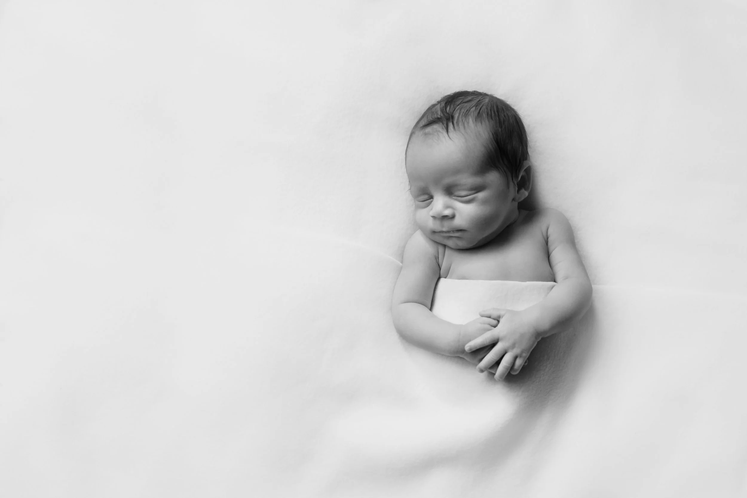 Black and white photo of a sleeping newborn baby with closed eyes, lying on its side, wrapped in a blanket, on a soft surface.