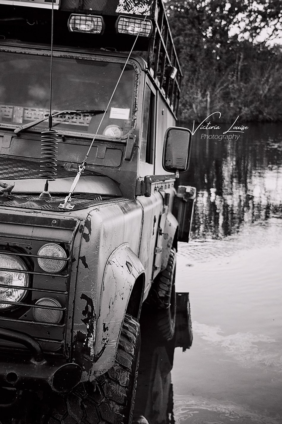A black and white photo of a rugged off-road vehicle parked by a body of water with trees reflected in the water. The vehicle shows signs of wear and has various antennas and equipment mounted on it.