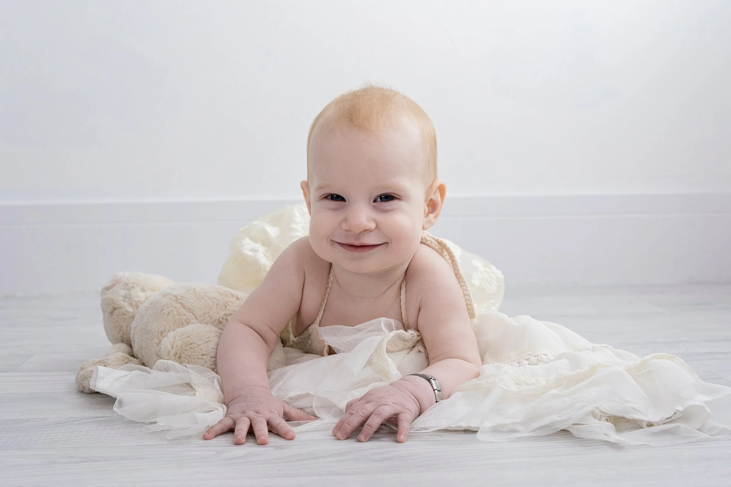 Smiling baby with light hair, lying on the floor, surrounded by plush toys and dressed in light, airy clothing.