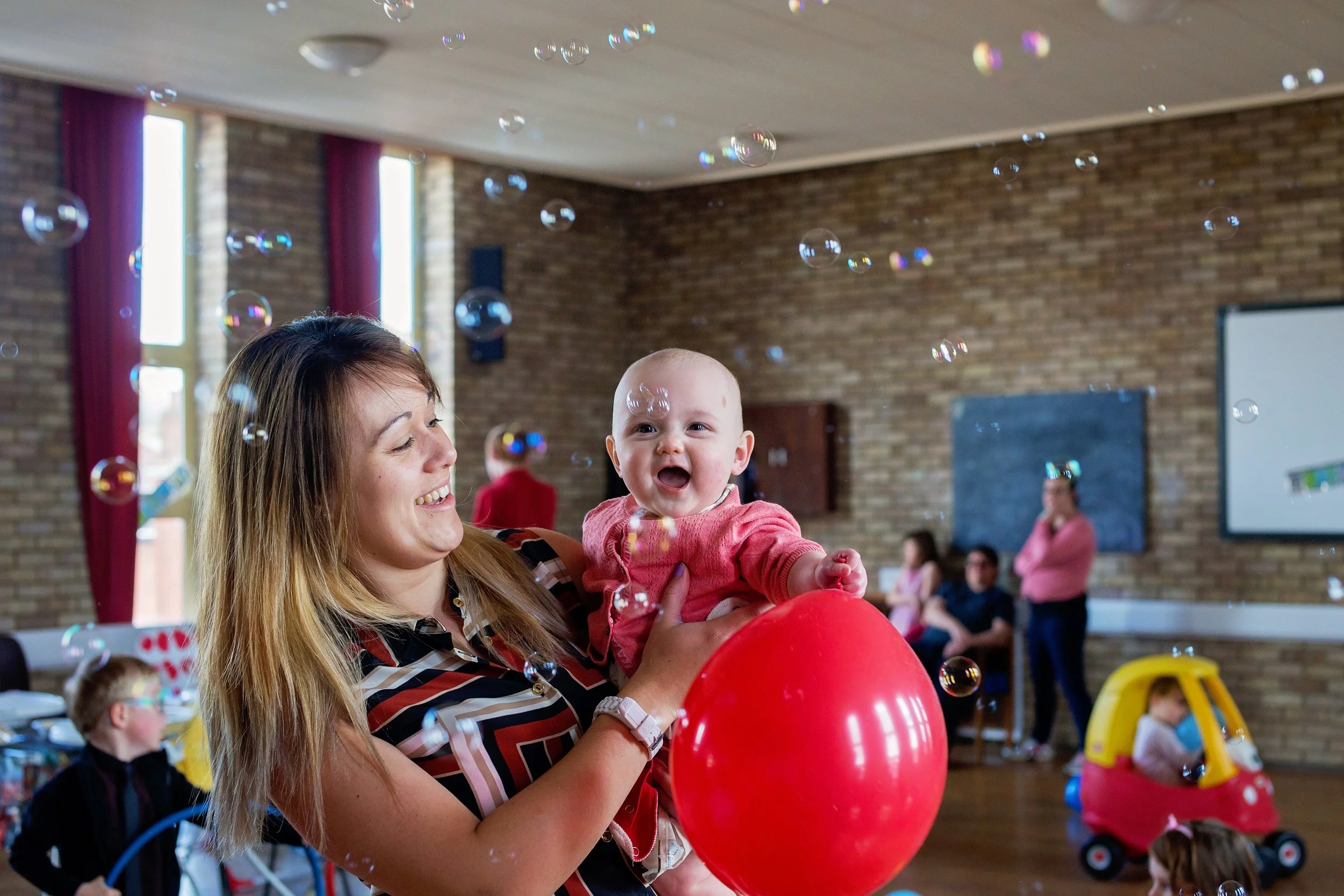 A woman holding a smiling baby in a pink shirt at an indoor children's party, with bubbles floating in the air, and children playing with toys in the background.