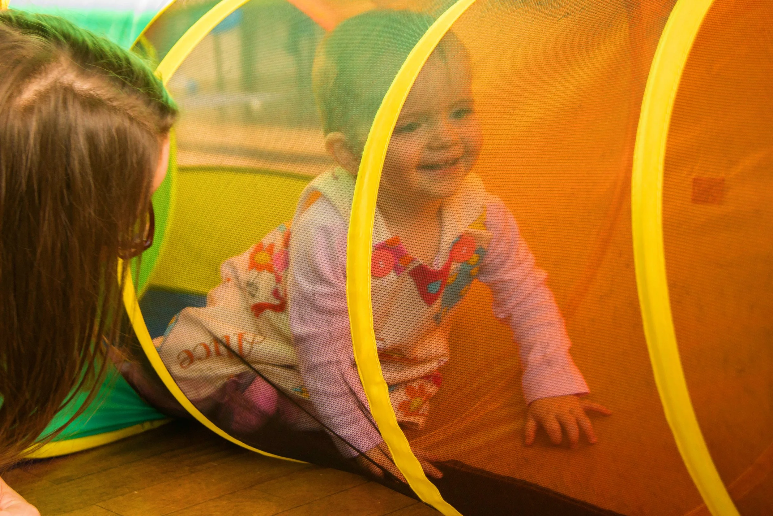 A young child crawling through a colorful play tunnel with an adult observing.