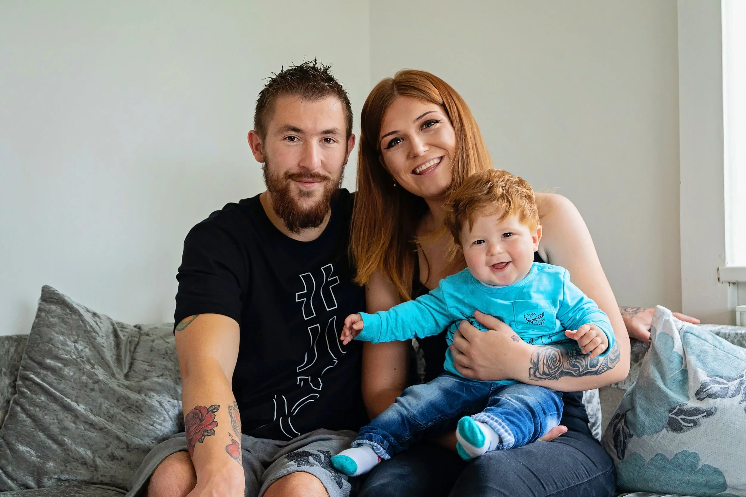A happy family of three sitting on a sofa inside a home, smiling at the camera. The family includes a man with a beard and tattoos, a woman with red hair, and a baby boy with red hair wearing a blue shirt.
