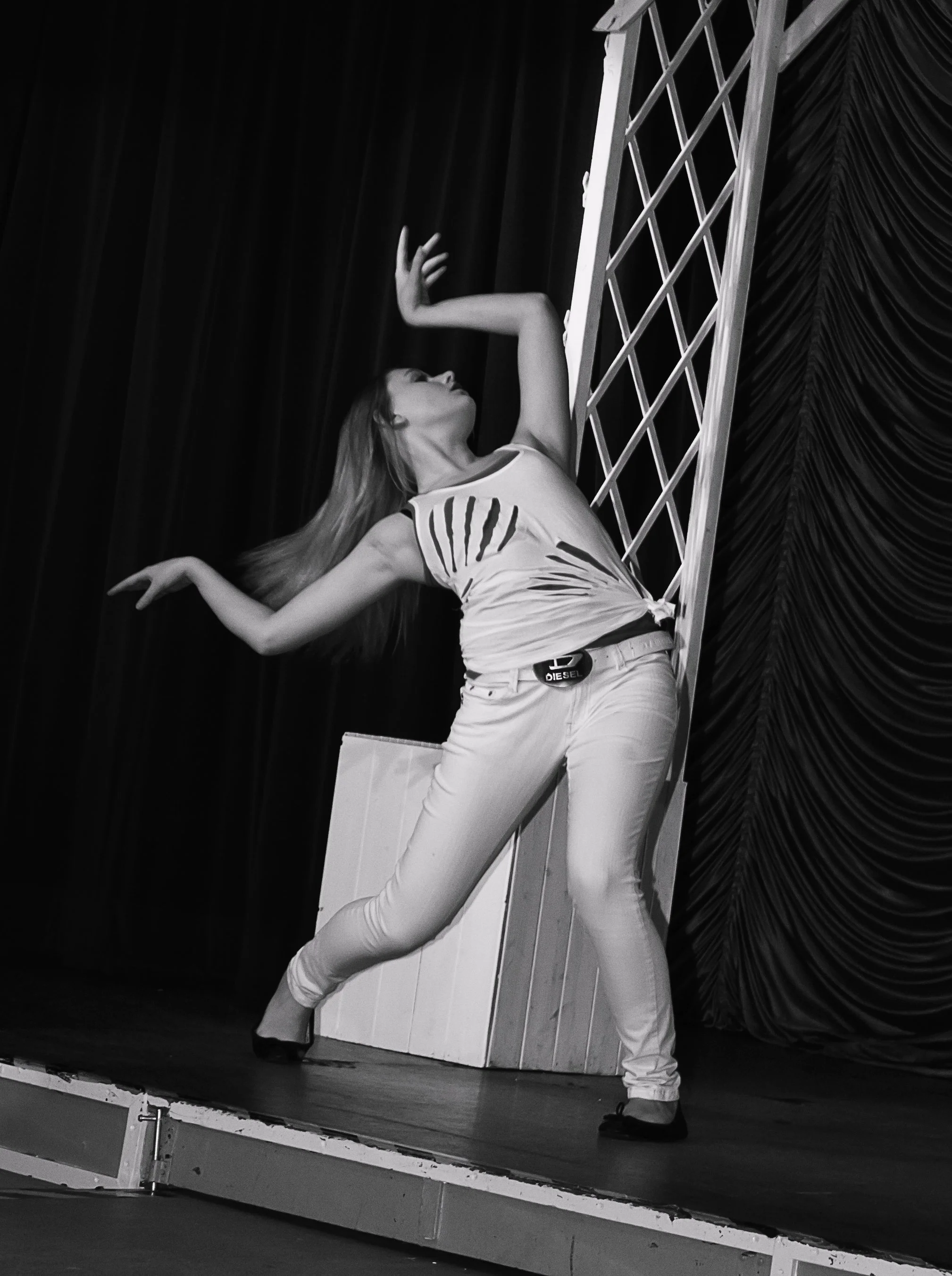 A woman dancing on stage, leaning back with arms raised and hair flowing, wearing light-colored jeans and a sleeveless top, with a stage curtain and decorative backdrop in the background.