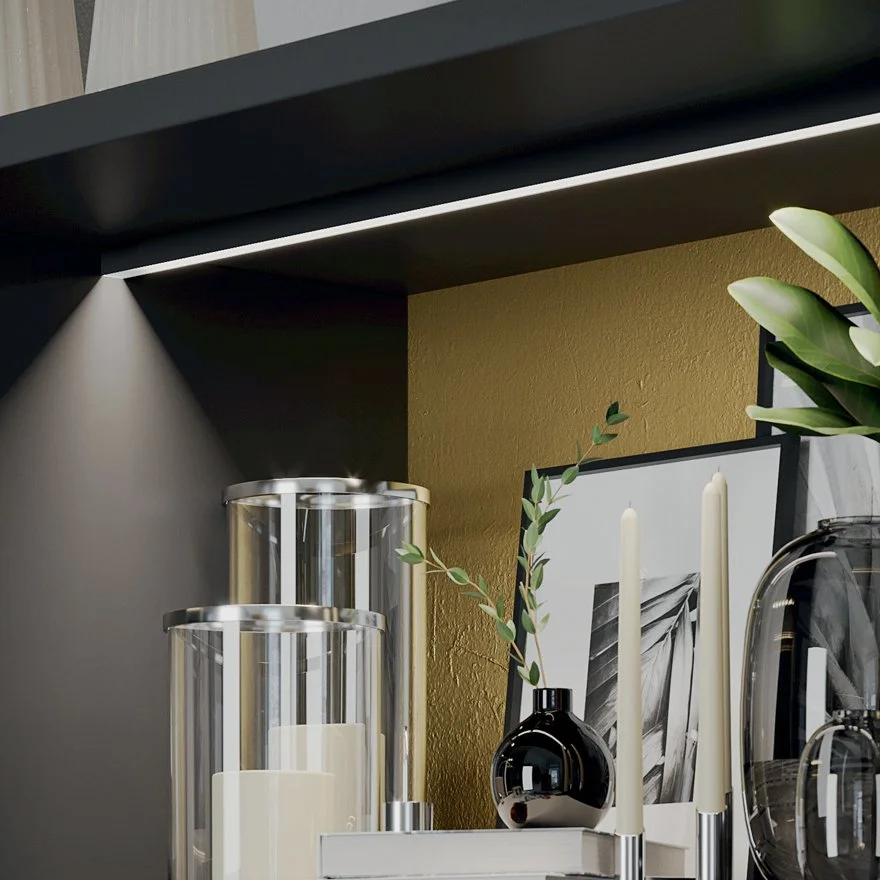 Modern interior shelf decor with glass candle holders, black vase, framed photos, and green leaves on a gold background, with recessed lighting.