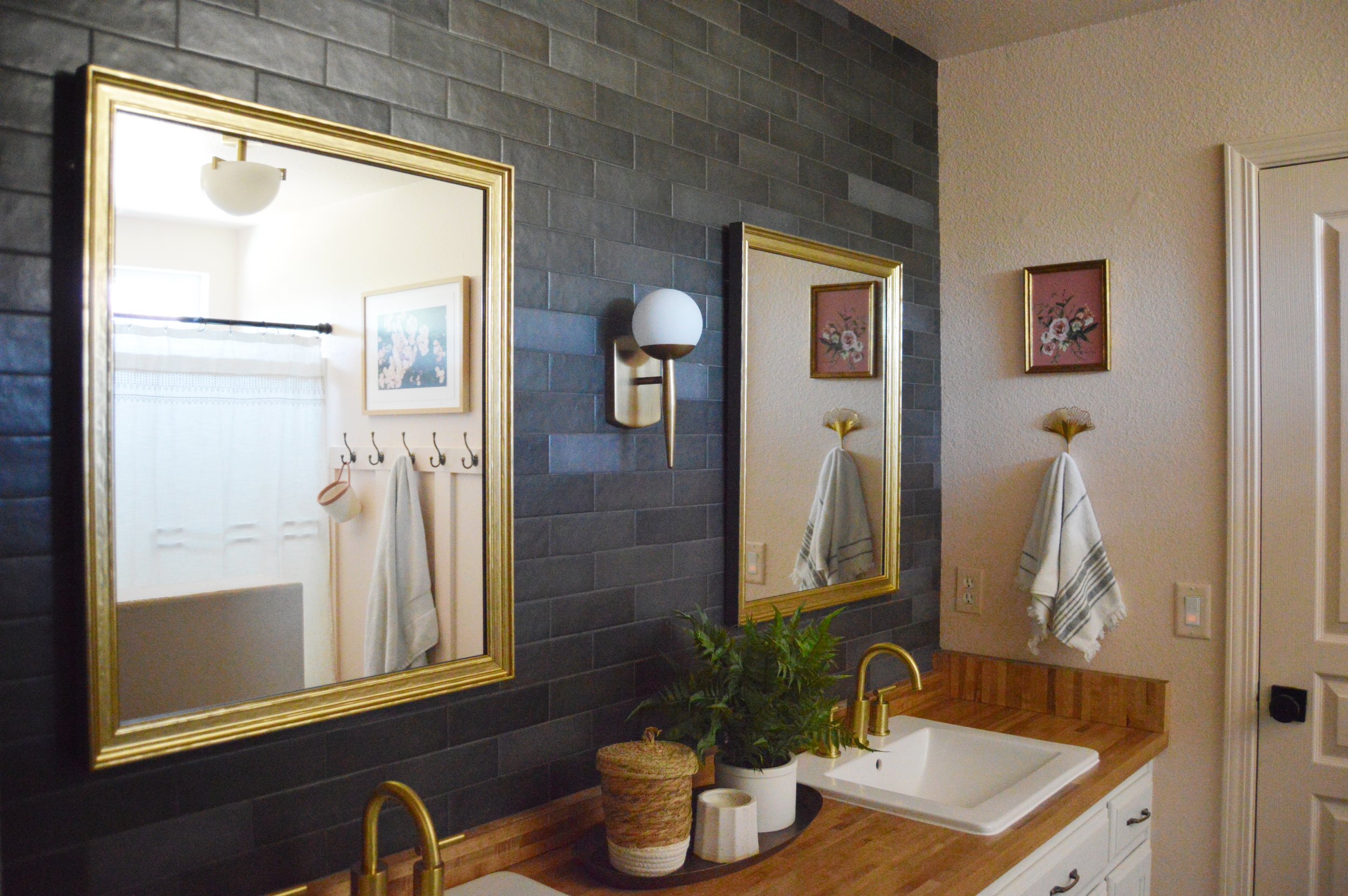 Bathroom interior with two gold-framed mirrors, a wooden countertop, two white sinks with brass faucets, a wall-mounted light fixture, decorative plants, and white towels.