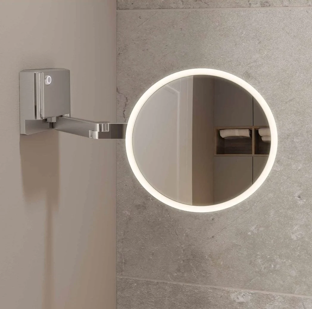 Wall-mounted round vanity mirror with LED light and adjustable arm on a tiled bathroom wall.