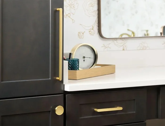 Bathroom countertop with a wooden tray holding an alarm clock and a container, beside dark cabinets with gold handles.