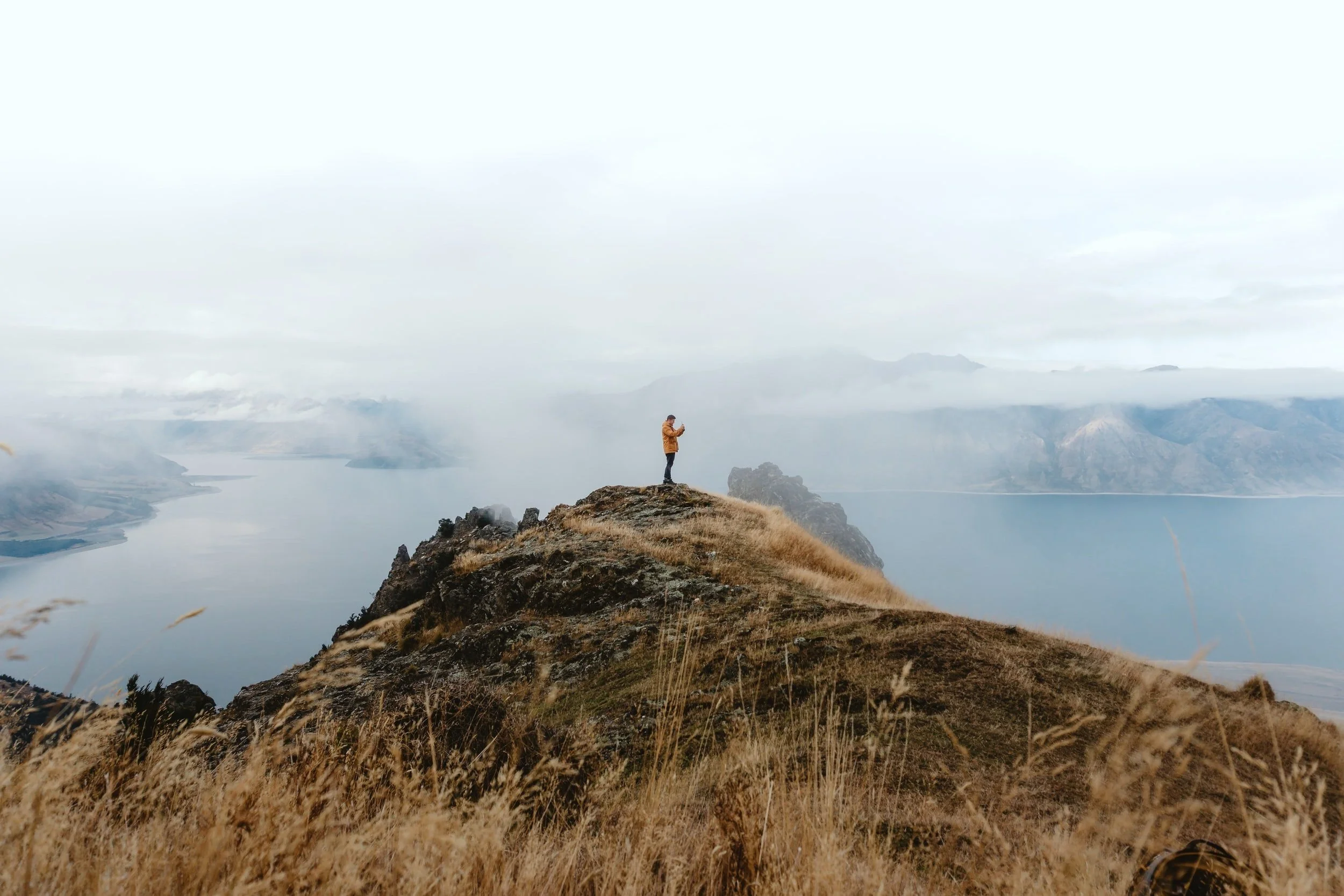 Person standing on a mountain ridge overlooking a lake and distant mountains.