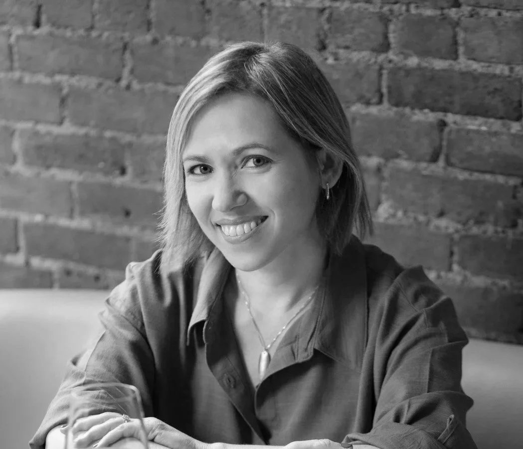 A woman with shoulder-length hair smiling while sitting at a table with a glass of wine, with a brick wall in the background.