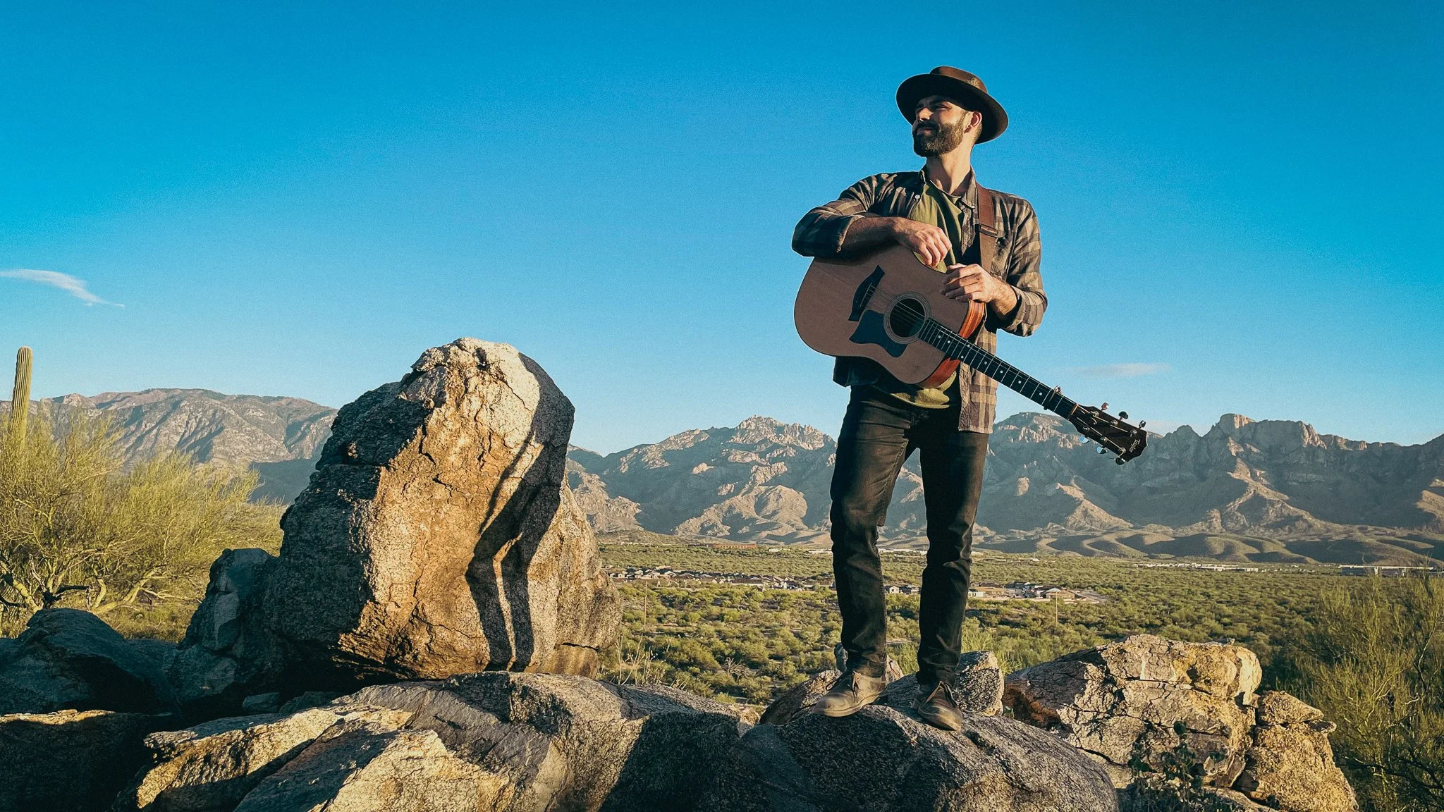 A man standing on rocks outdoors in a desert landscape, holding an acoustic guitar and wearing a wide-brimmed hat. Mountains and a clear blue sky are in the background.