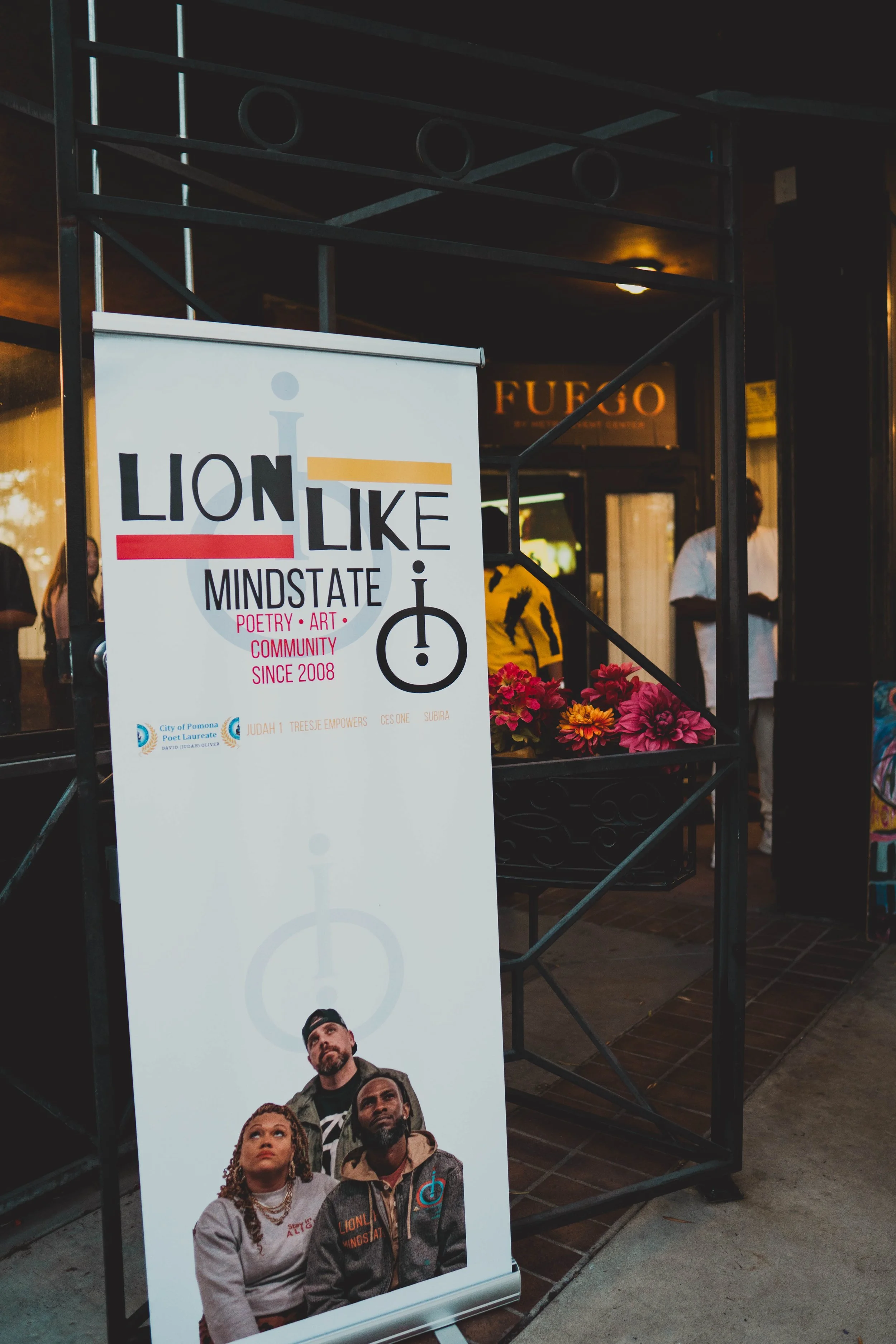 A large vertical banner outside a building with a sign that reads "LION LIKE MINDSTATE" and text indicating poetry, art, and community since 2008. The banner features an image of three people looking upward.