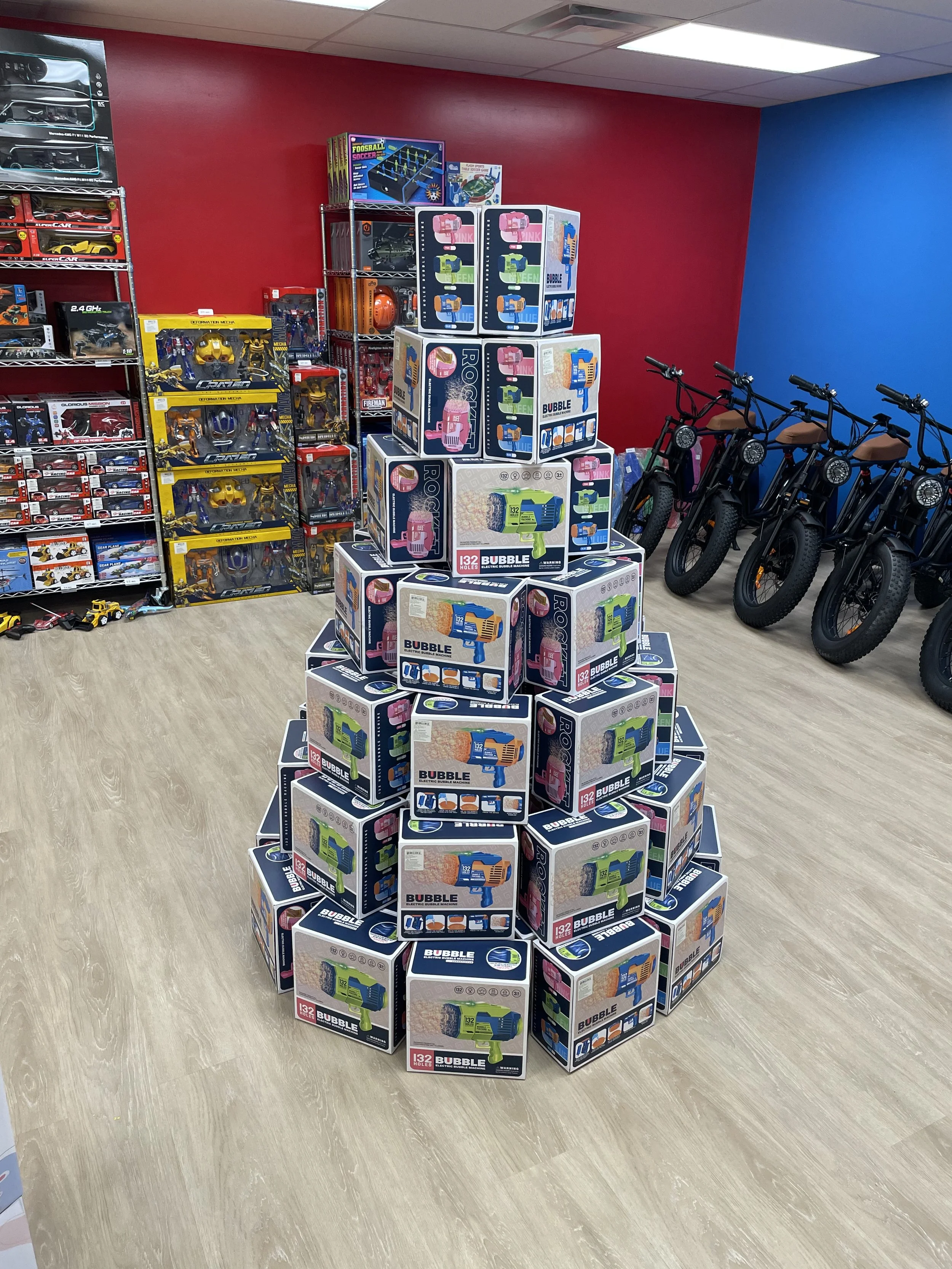 Display of stacked boxes of bubble guns in a toy store, with shelves of toy cars and remote-controlled vehicles on the left, and bikes lined up against a blue wall on the right.