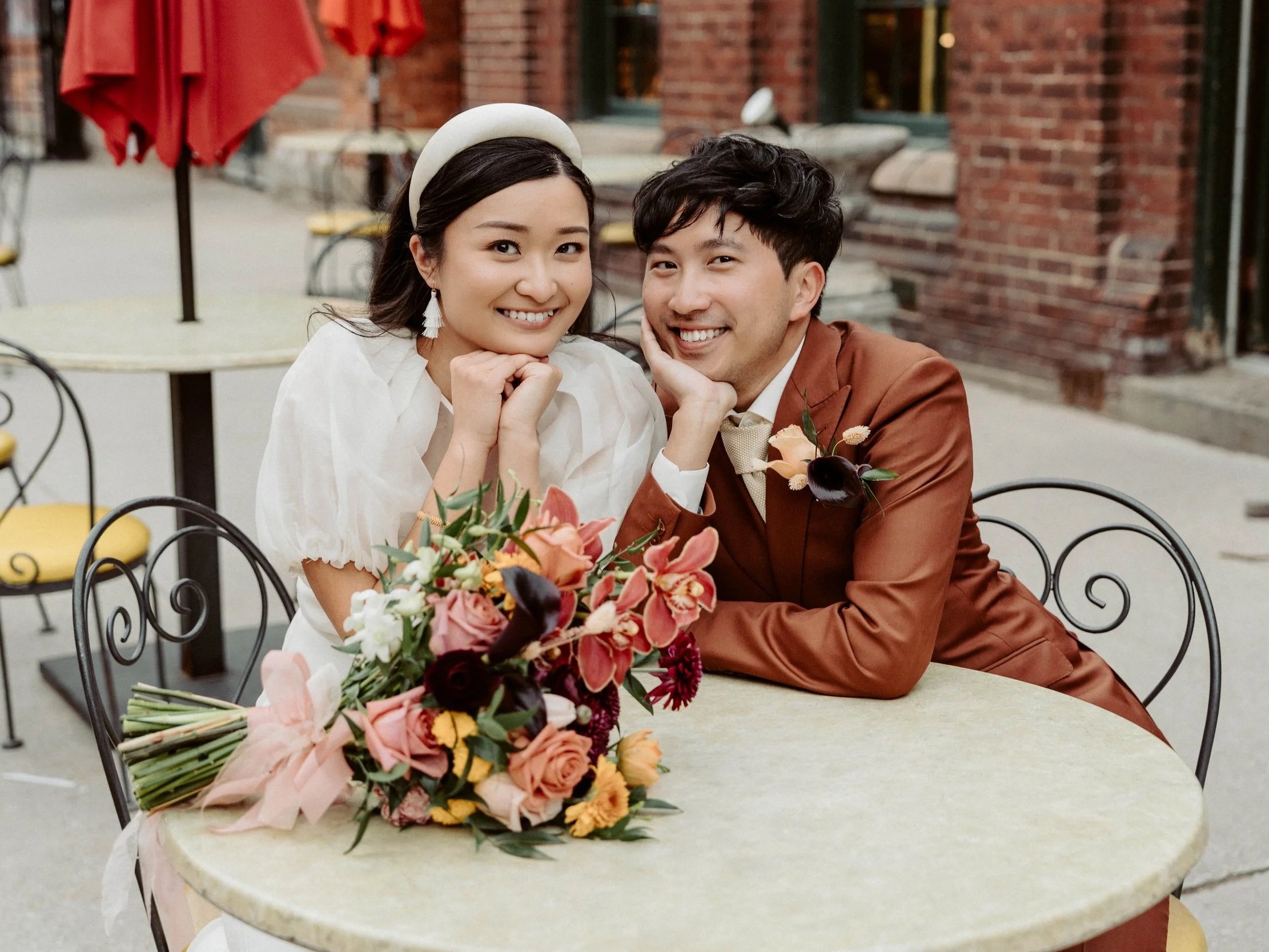 A smiling woman and man sitting at a table outdoors with a large floral bouquet in front of them. The woman has dark hair with a white headband, and the man has dark, wavy hair and is wearing a brown suit. They are resting their chins on their hands 