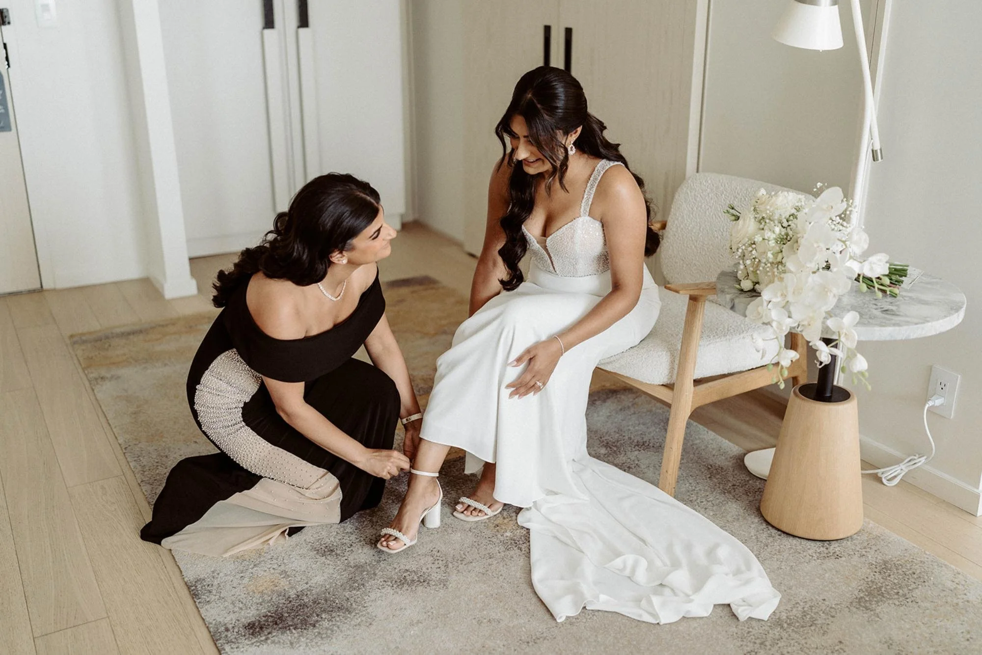Bride getting ready as another woman kneels to fasten her shoe in a bright modern room with white flowers on a side table.