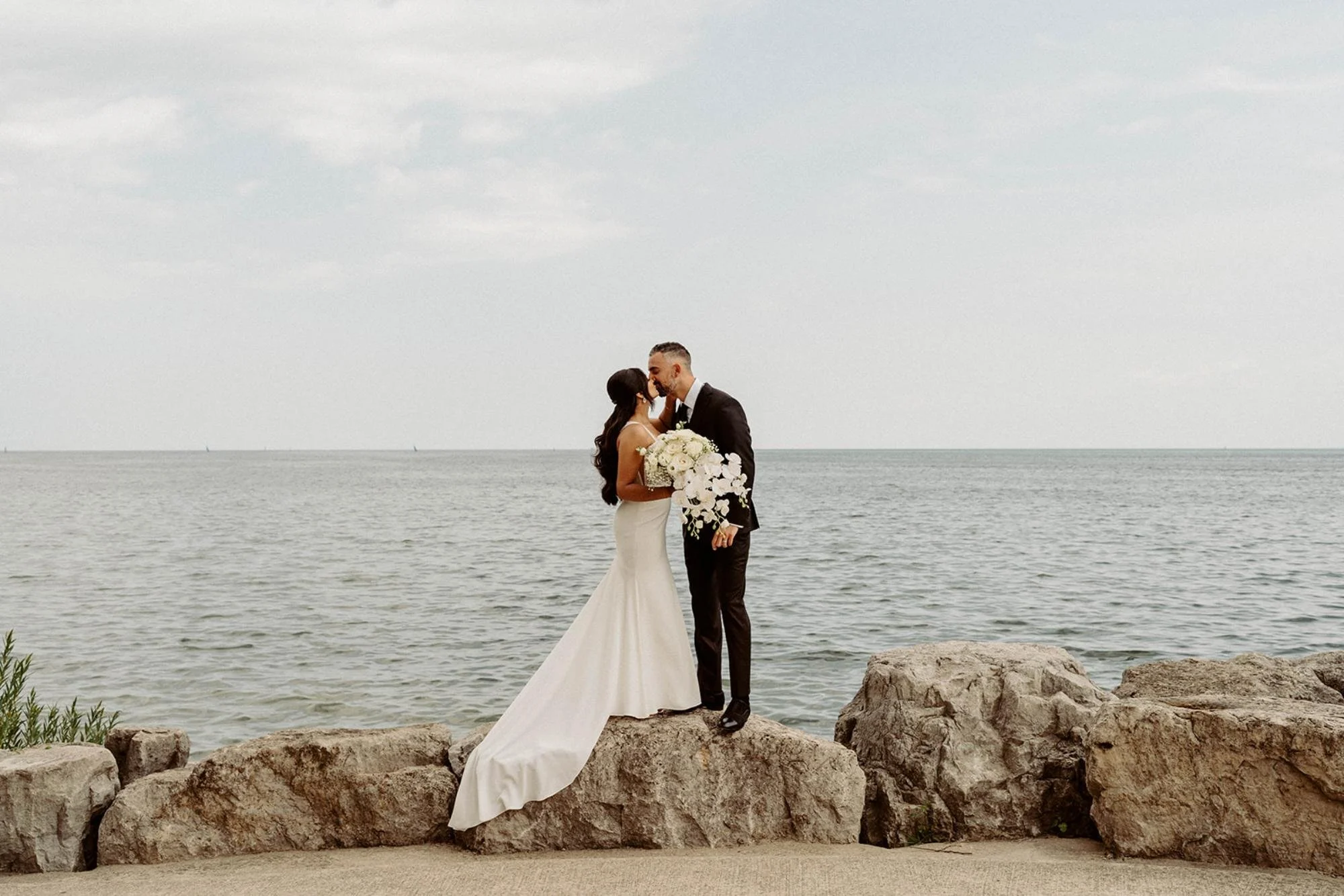 Bride and groom standing on large rocks by the water, sharing a kiss with the lake and sky in the background.