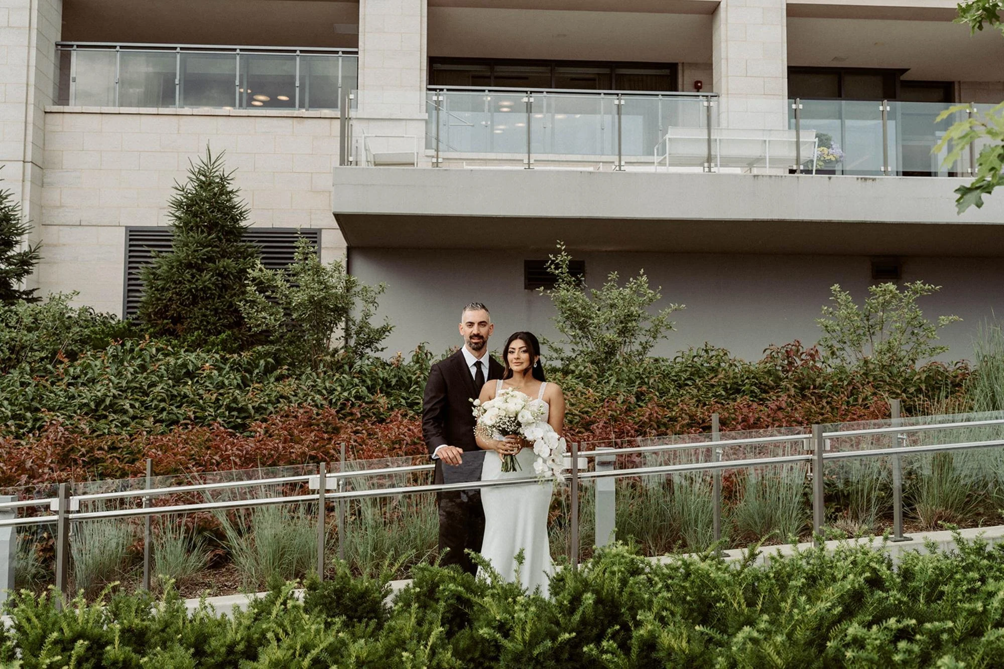 Bride and groom posing together in front of a landscaped garden area with shrubs and a modern building behind them.