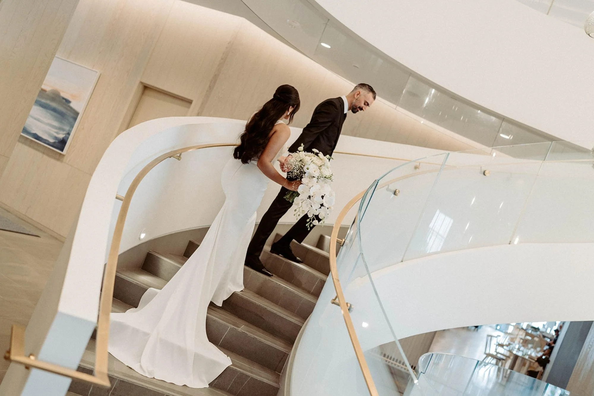 Bride and groom walking up a modern curved staircase, with the bride holding a white bouquet and her dress train trailing behind her.