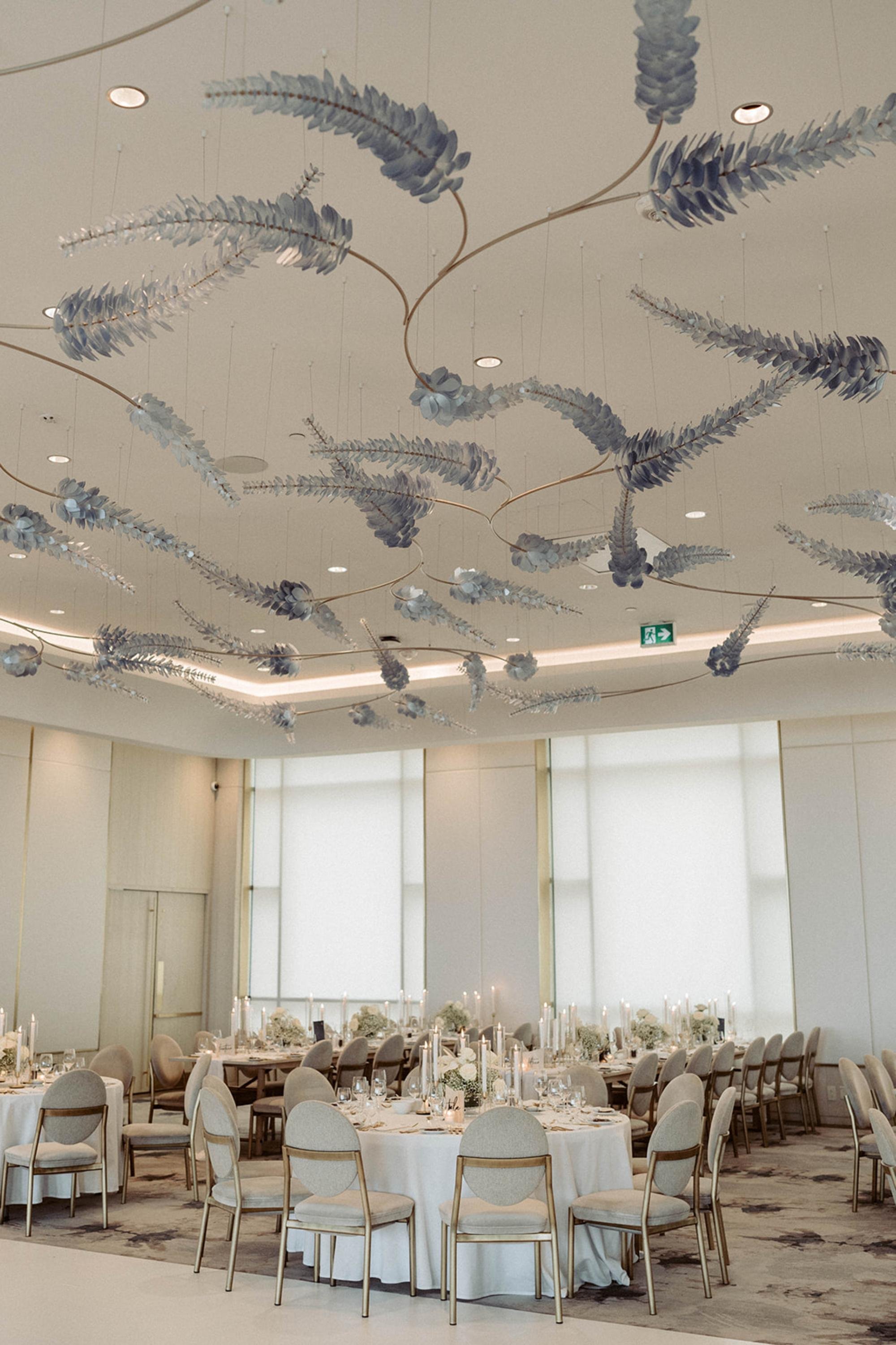 Wedding reception room with round tables, tall candles, white floral centerpieces, and sculptural hanging ceiling décor.