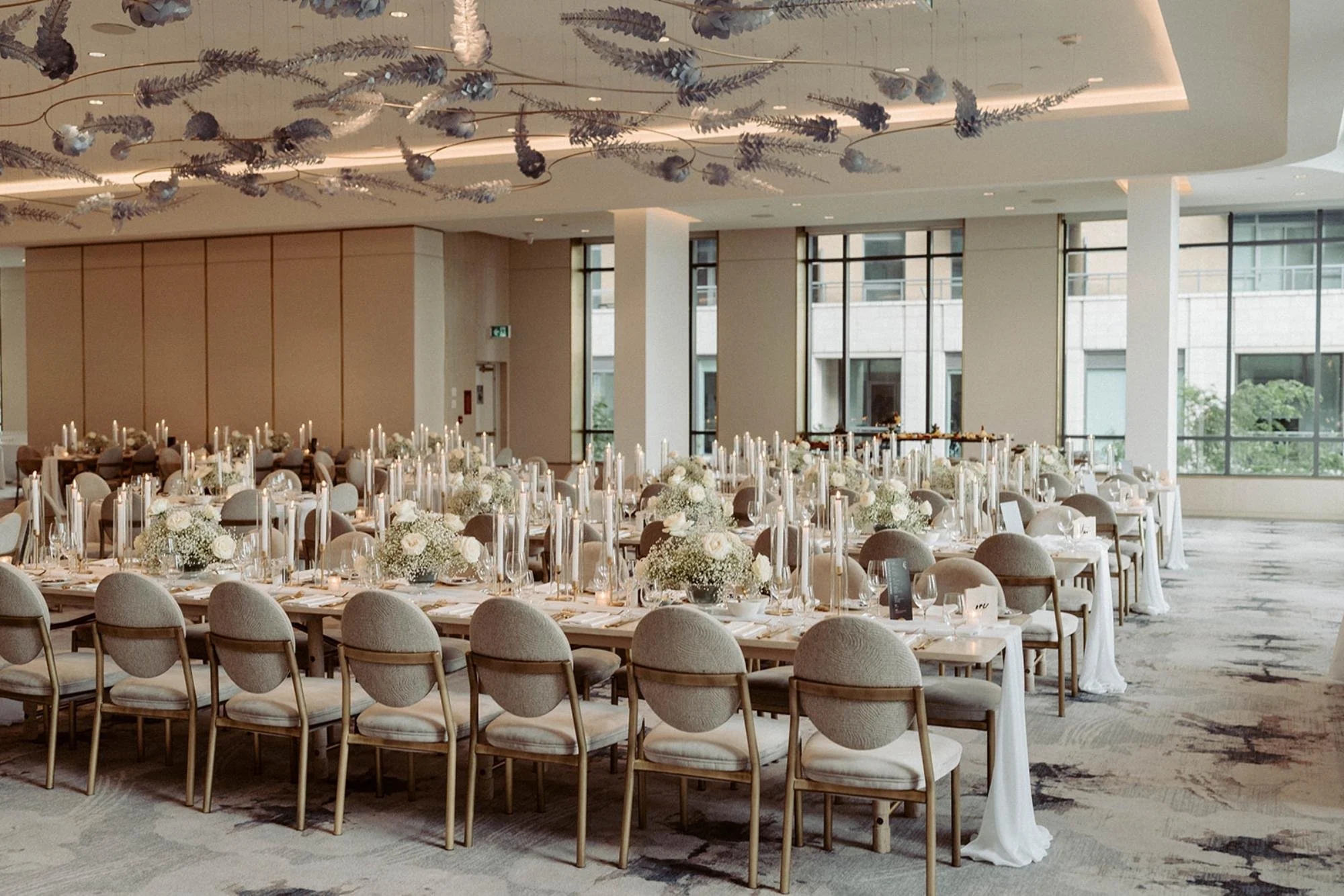 Long reception tables decorated with candles and white flowers beneath an artistic ceiling installation in a bright modern venue.