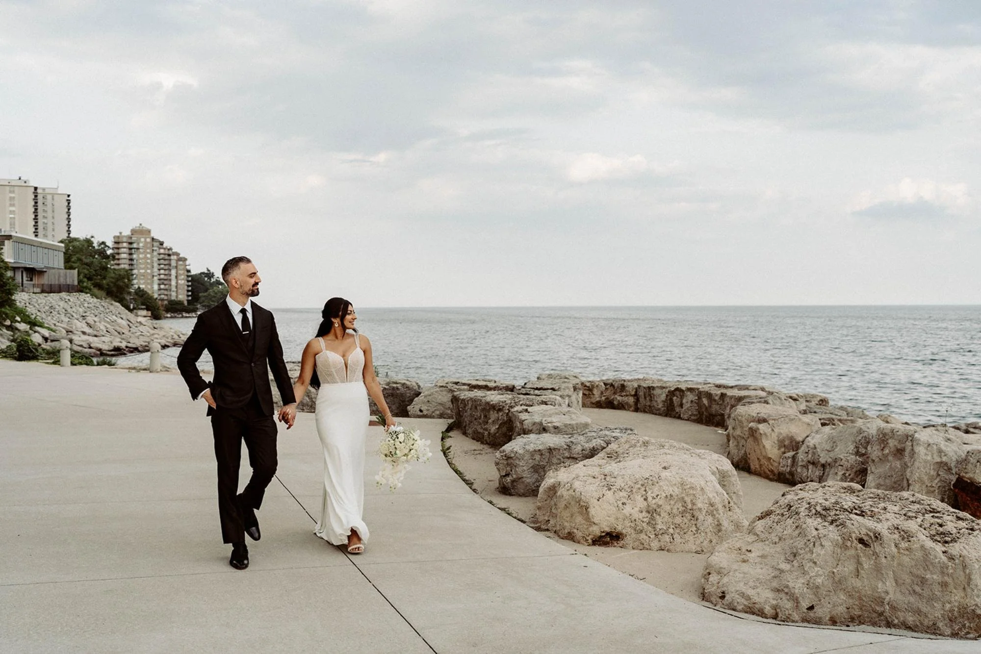 Bride and groom walking along a waterfront pathway holding hands, with large rocks and lake views beside them.